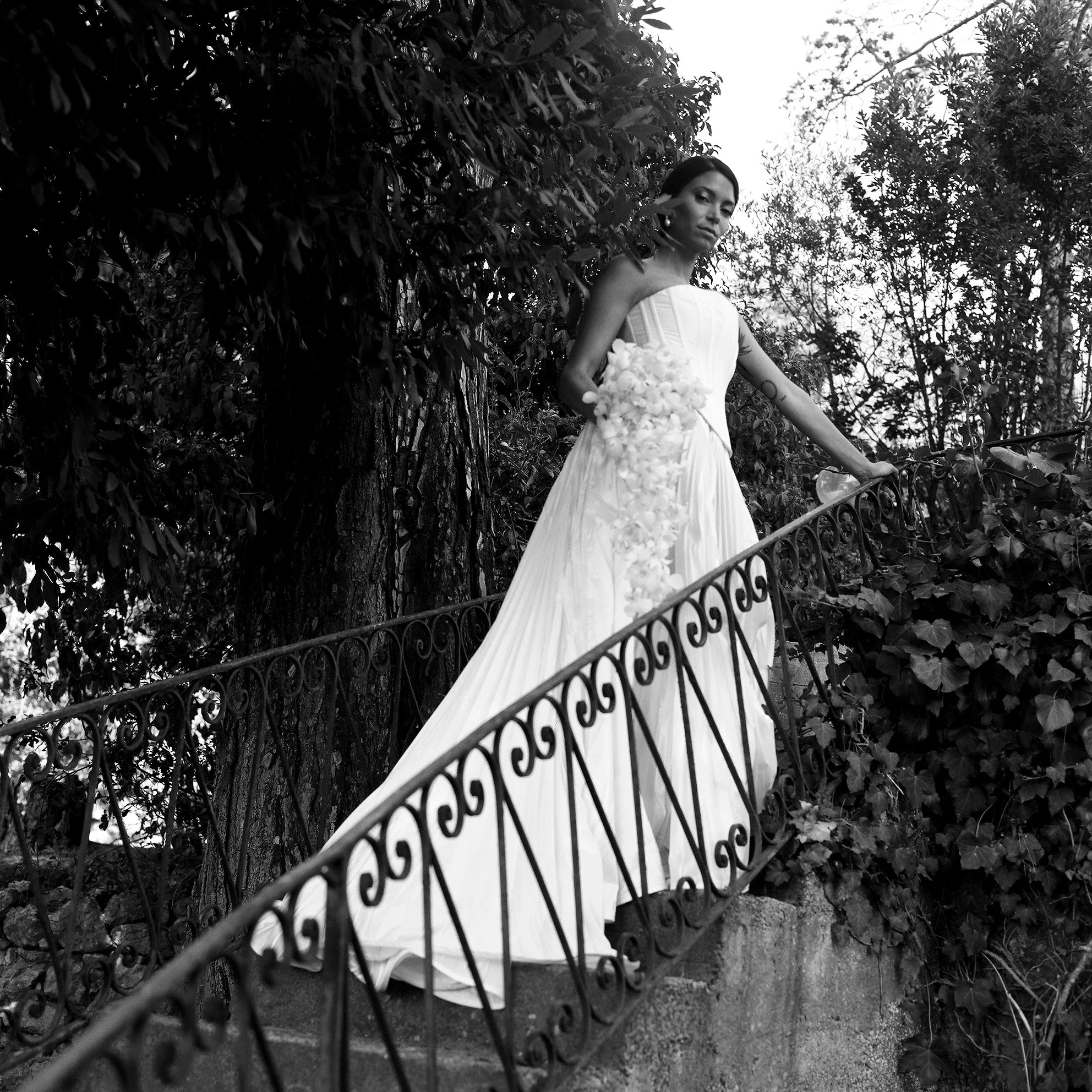 Donna in abito da sposa che si tiene alla ringhiera di una scalinata, circondata da alberi e vegetazione, in una scena in bianco e nero.