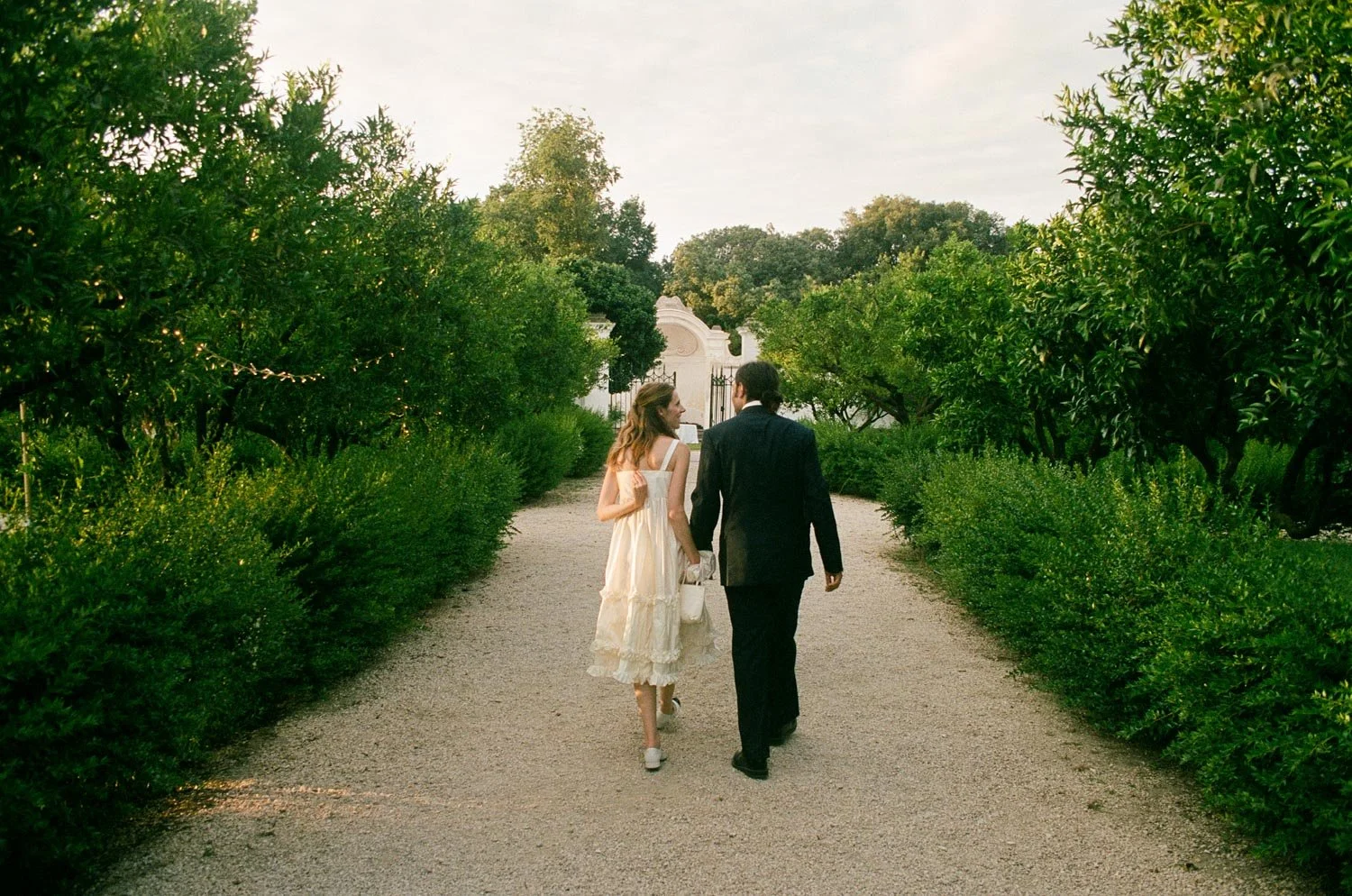 Una coppia di persone vestite elegantemente cammina lungo un sentiero circondato da cespugli e alberi verdi, con un cancello e edifici bianchi sullo sfondo.