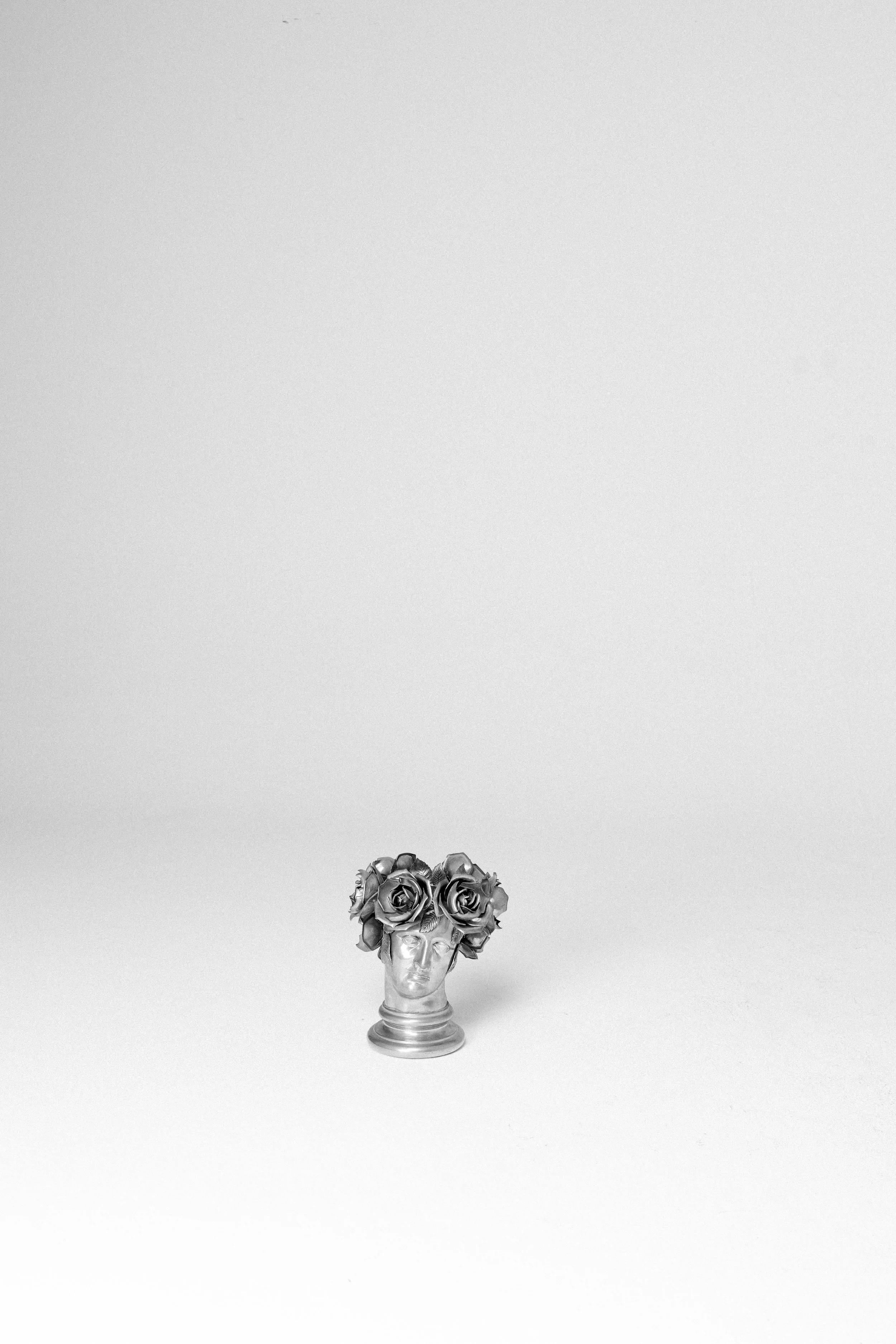 Statua di una testa umana con un'acconciatura fatta di rose, di colore argento, posizionata su uno sfondo bianco