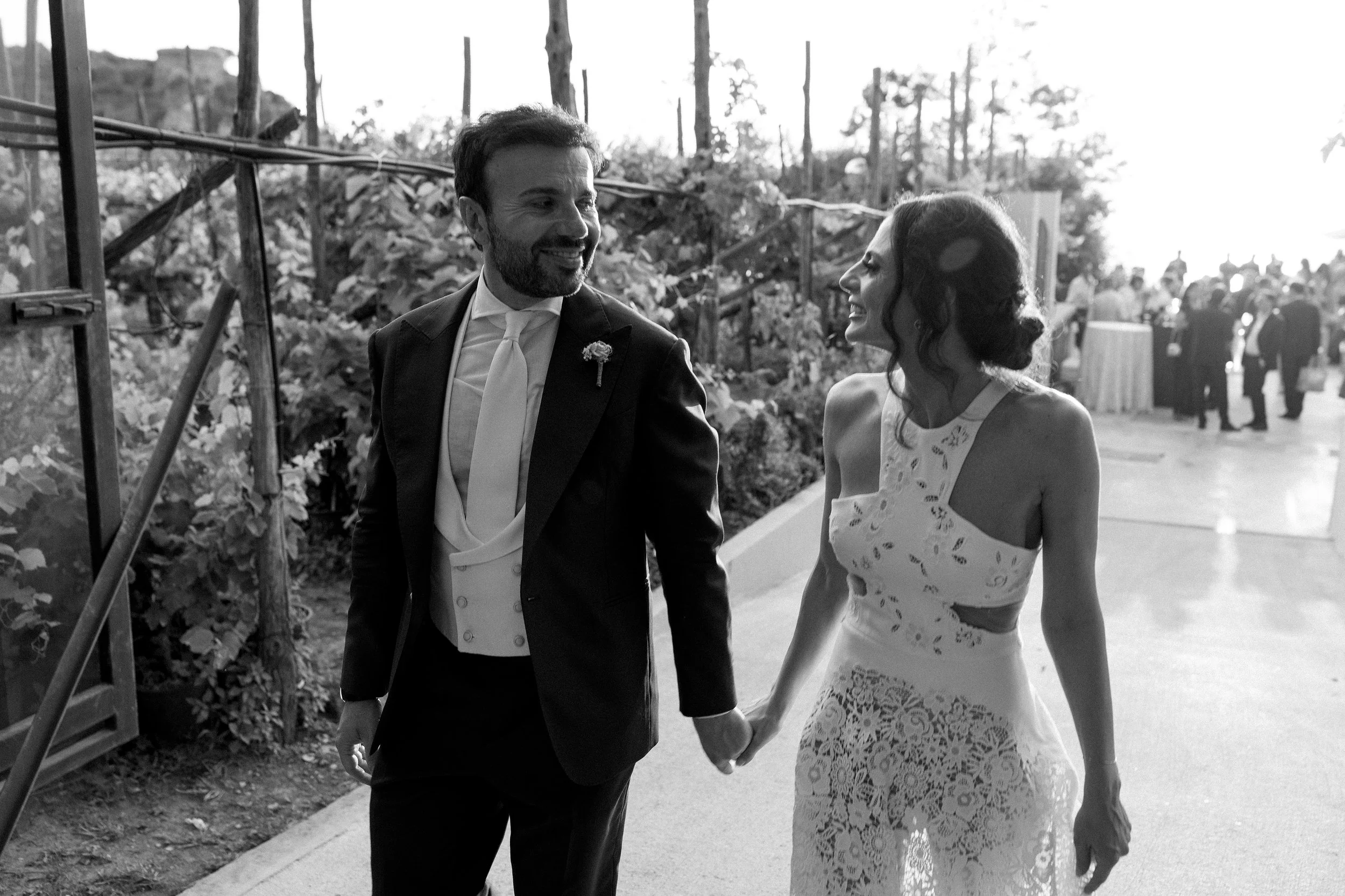 Due persone, un uomo e una donna, si tengono per mano e sorridono, probabilmente in un evento speciale come un matrimonio, all'aperto con un gruppo di persone sullo sfondo.