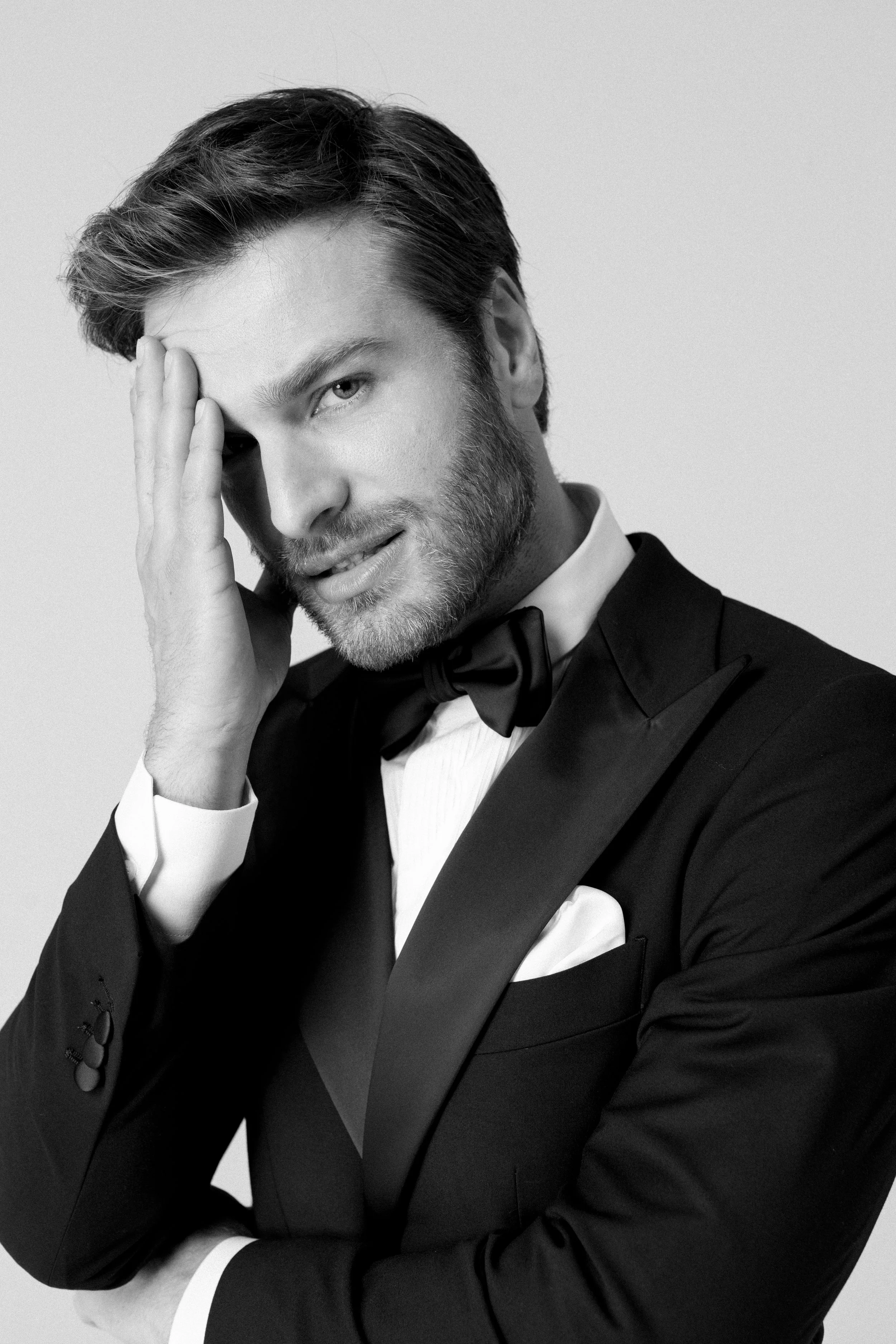 Ritratto in bianco e nero di un uomo con abito nero, camicia bianca e papillon, che tocca la tempia con la mano destra, con espressione sicura.