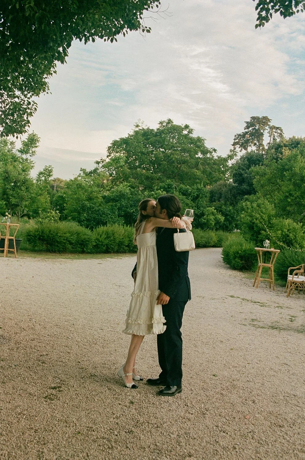 Una coppia si abbraccia e si bacia in un giardino con alberi e sedie di legno all'aperto, sotto un cielo leggermente nuvoloso.