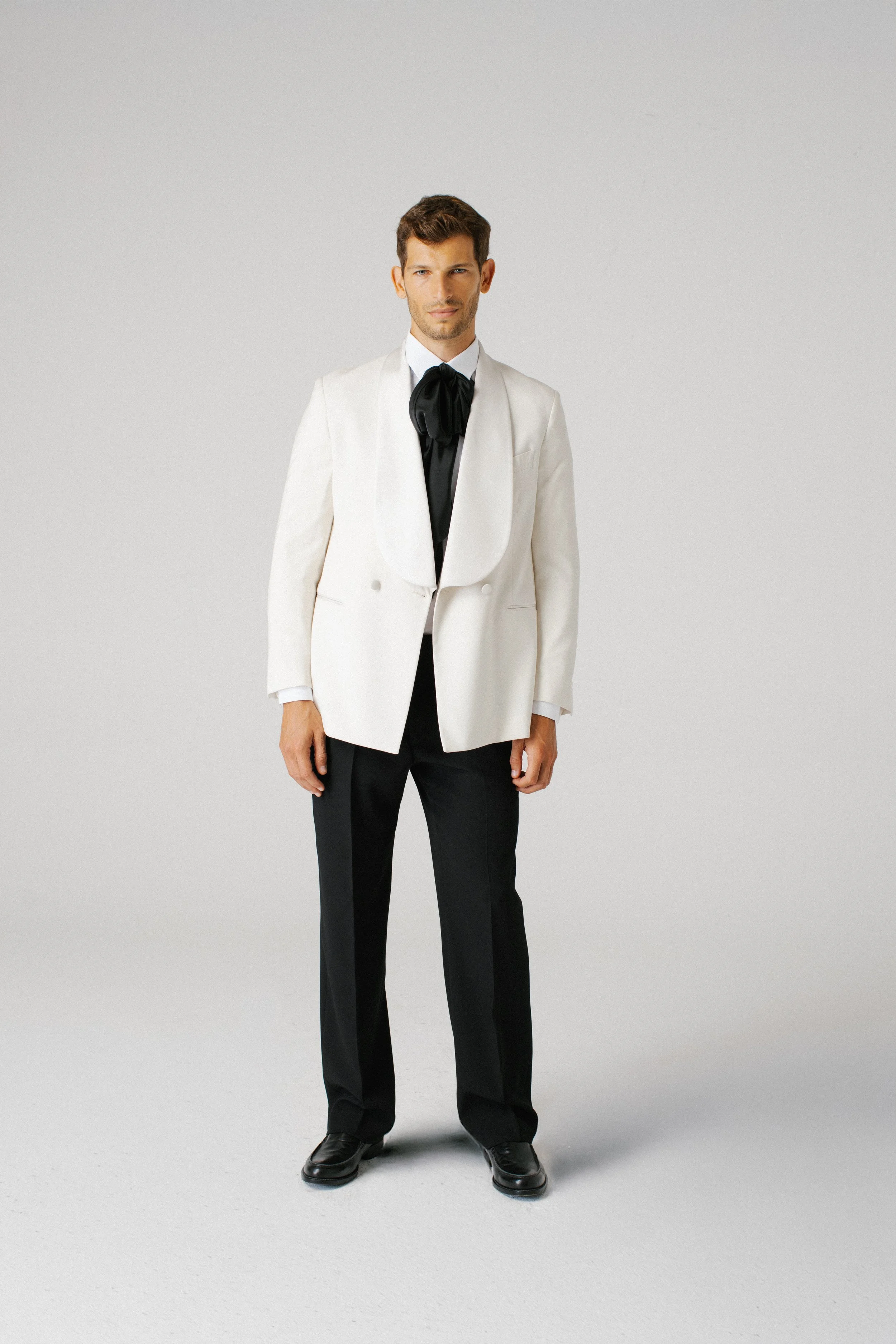 Uomo in abito nero e giacca bianca con cravatta nera, in piedi davanti a uno sfondo grigio chiaro.