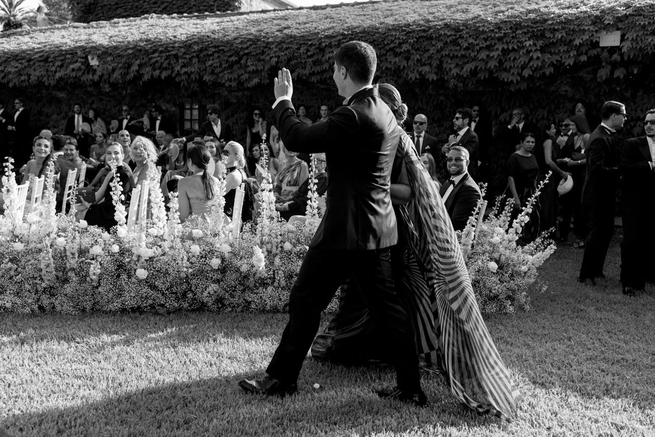 Una coppia in abito elegante balla in un giardino durante un matrimonio, con molti ospiti seduti e in piedi che li osservano, decorazioni floreali e pareti ricoperte di foglie in sottofondo.