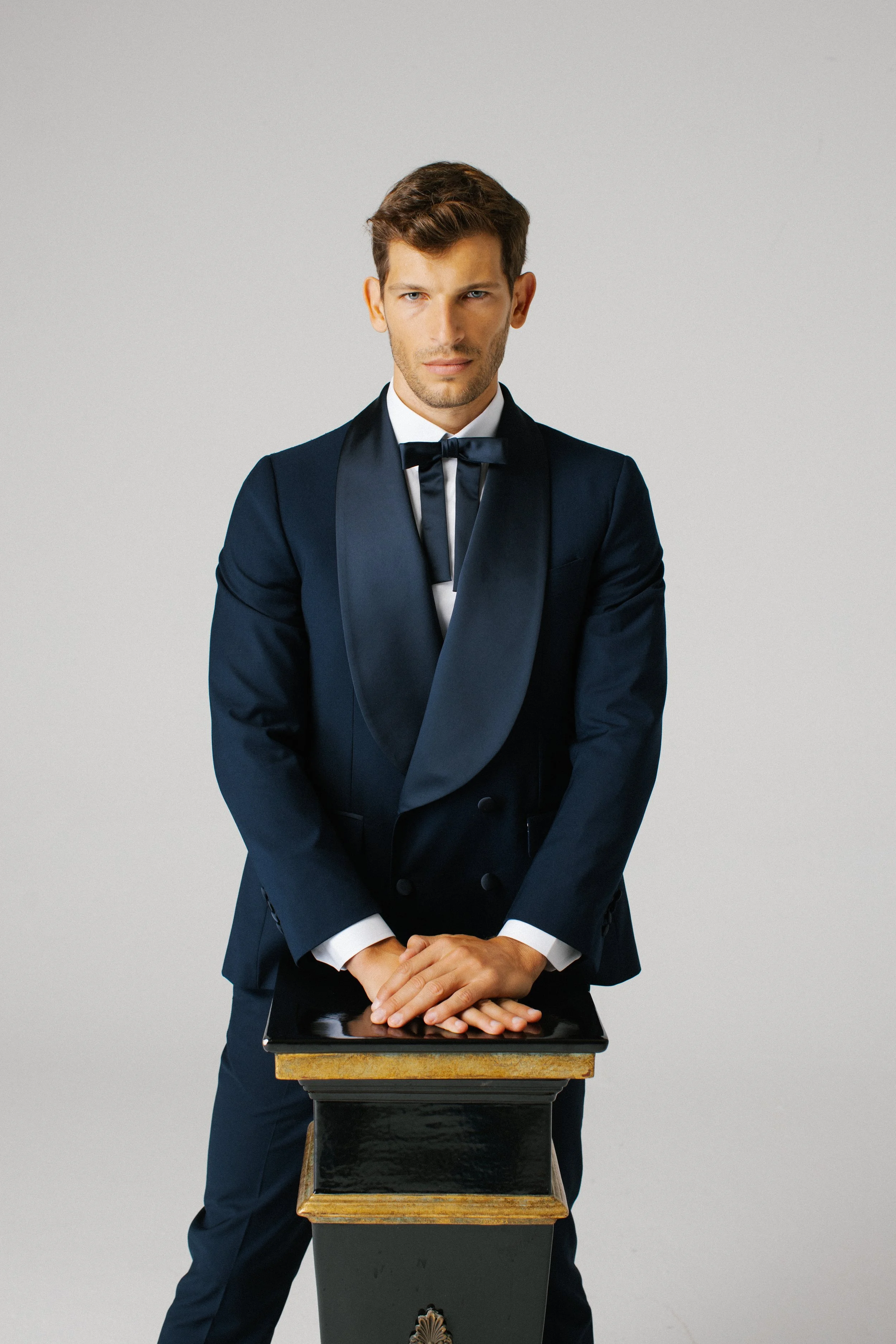 Uomo giovane in abito da sera elegante con a frill tra le mani, di fronte a uno sfondo grigio chiaro.