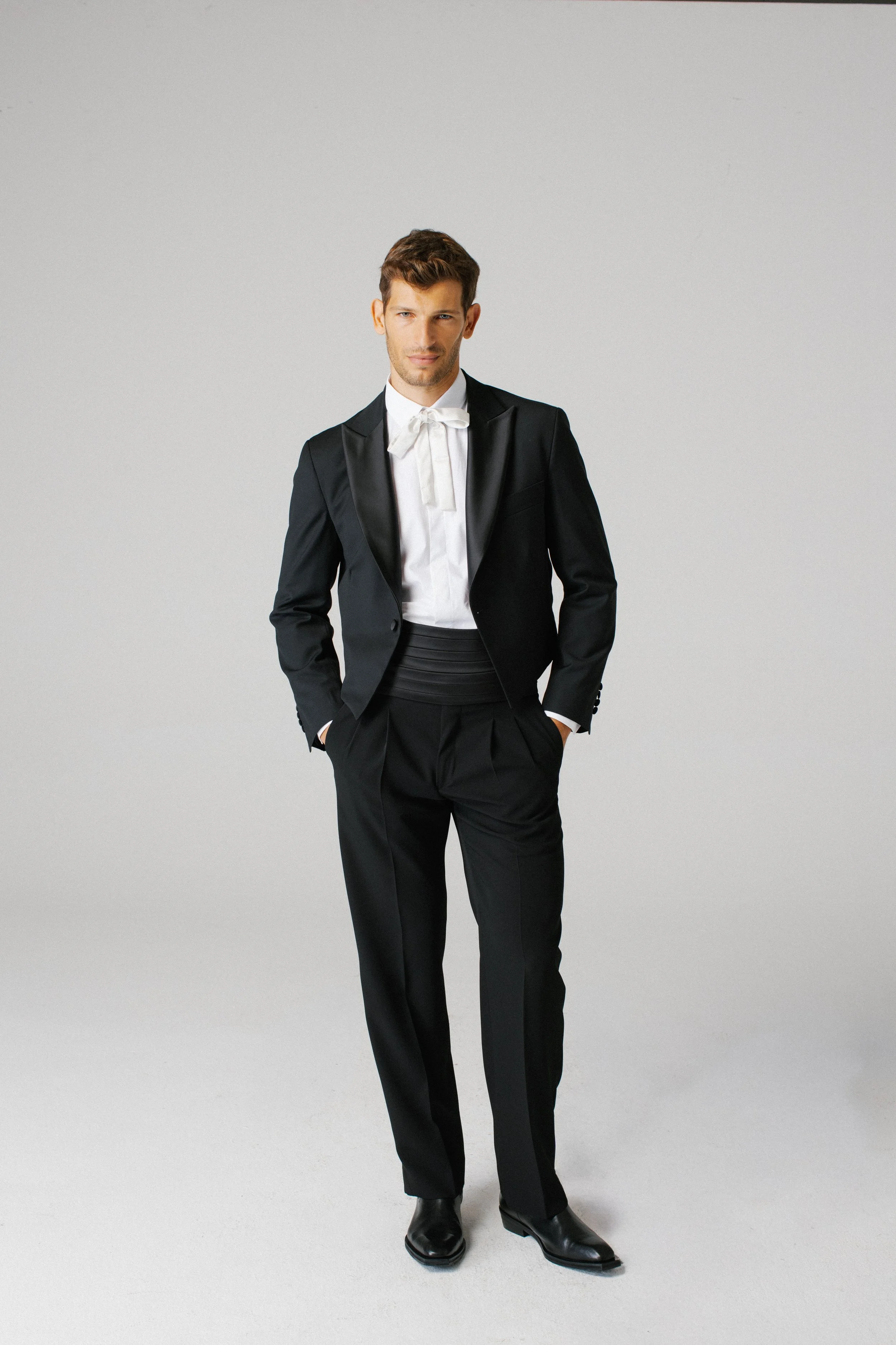 Uomo in abito nero con camicia bianca e cravatta in un ambiente neutro.