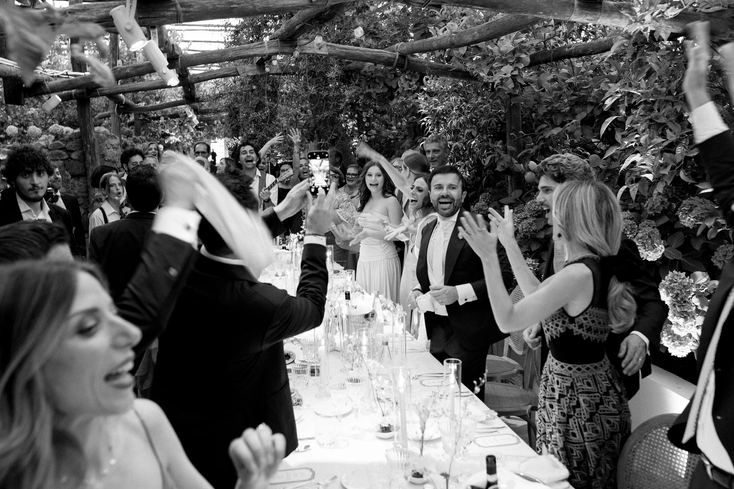 Una festa di matrimonio con persone che ridono, applaudono e si divertono, in un ambiente all'aperto con decorazioni floreali e luci sospese.