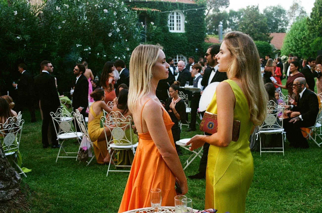 Due donne stanno chiacchierando in un giardino decorato per un evento sociale, con molte persone vestite elegantemente sullo sfondo.