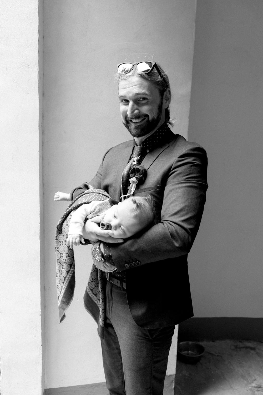 Uomo con giacca formale che tiene in braccio un neonato, entrambi sorridenti, in un'immagine in bianco e nero.