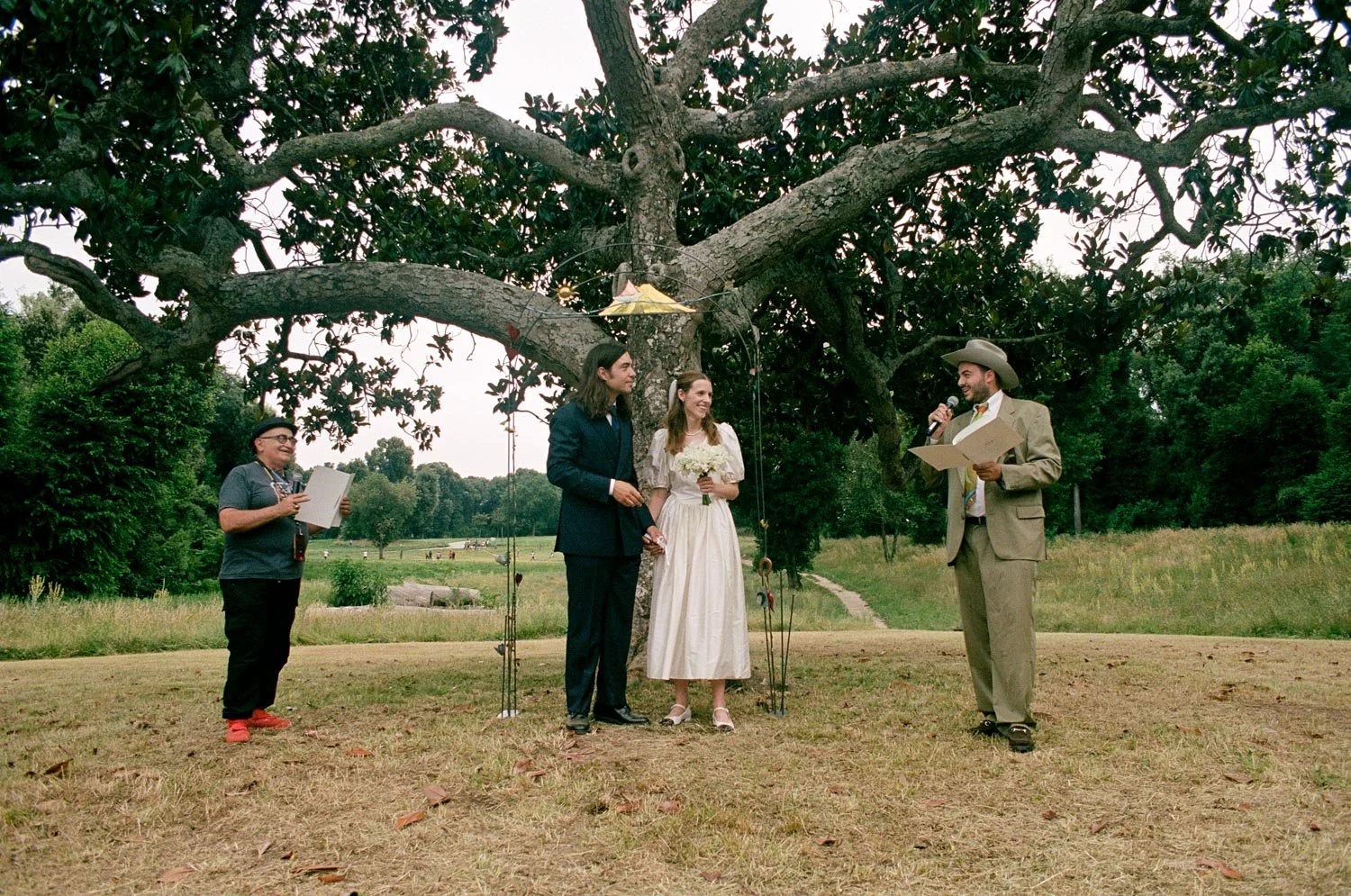 Uno sposo e una sposa che si tengono per mano durante una cerimonia di matrimonio all'aperto sotto un grande albero, con altre persone presenti, uno dei quali tiene un foglio e sta parlando.