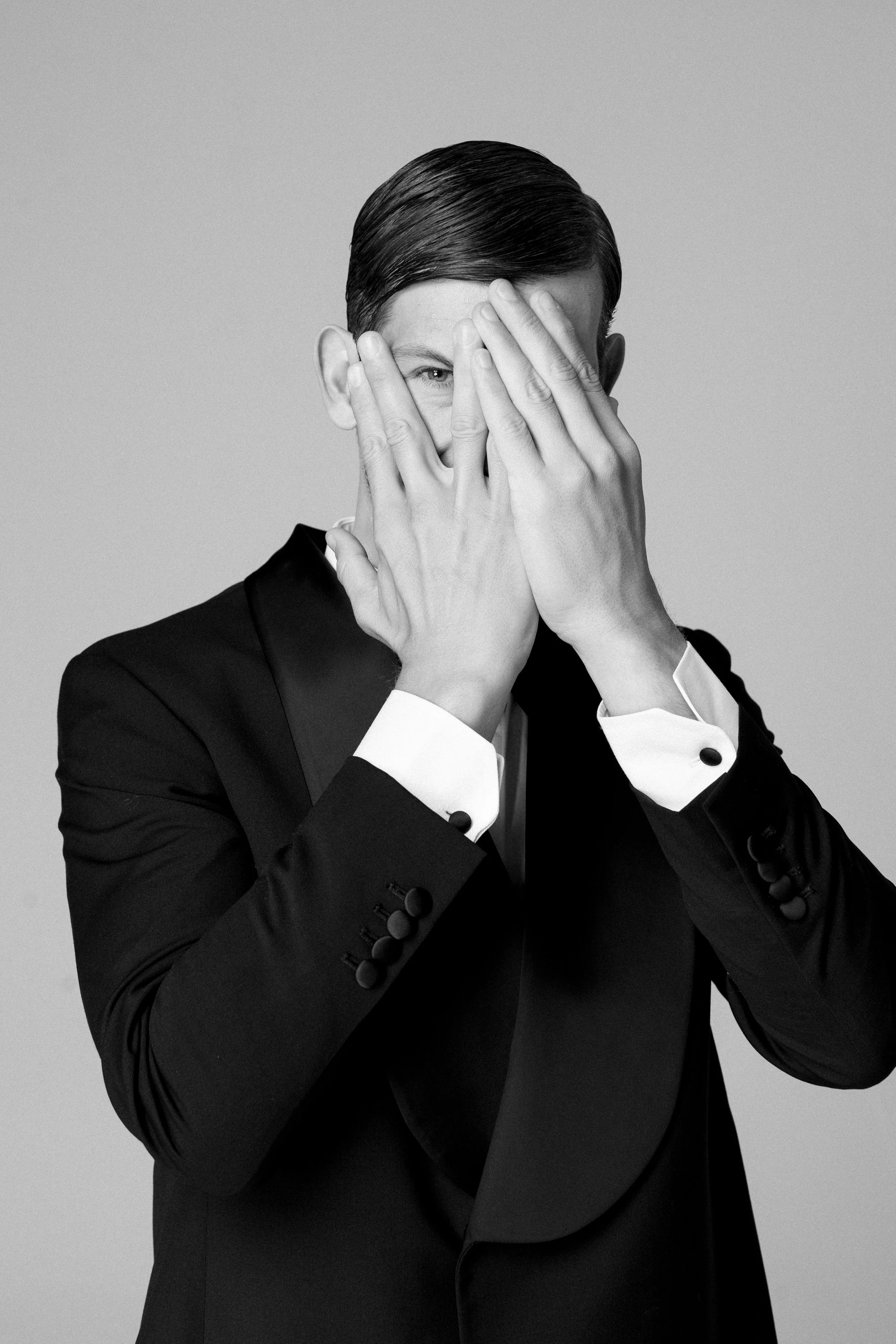 Uomo in abito nero con camicia bianca copre metà del volto con entrambe le mani.