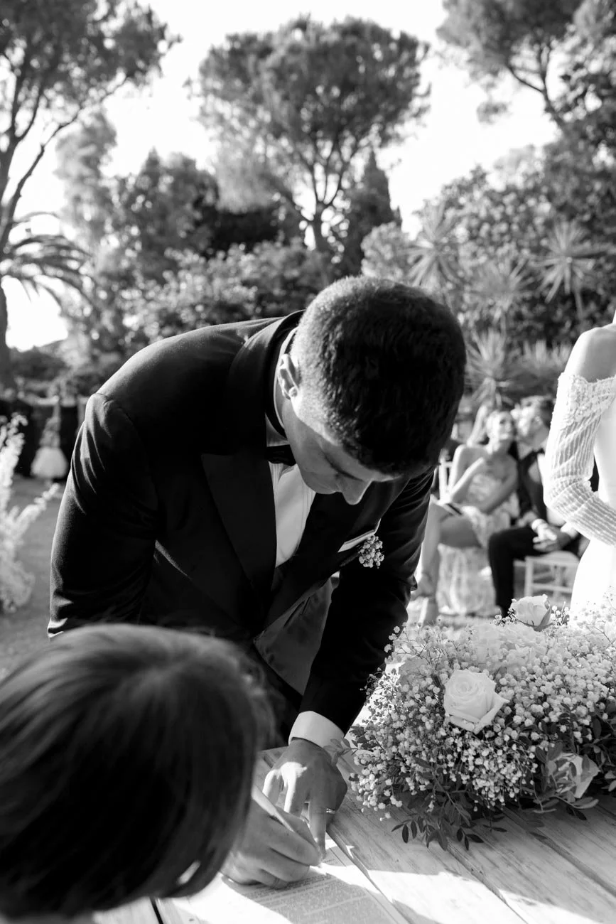 Un uomo in abito da cerimonia sta firmando un documento durante un matrimonio, con un bouquet di fiori su un tavolo accanto a lui, mentre altre persone sono presenti sullo sfondo in un giardino.