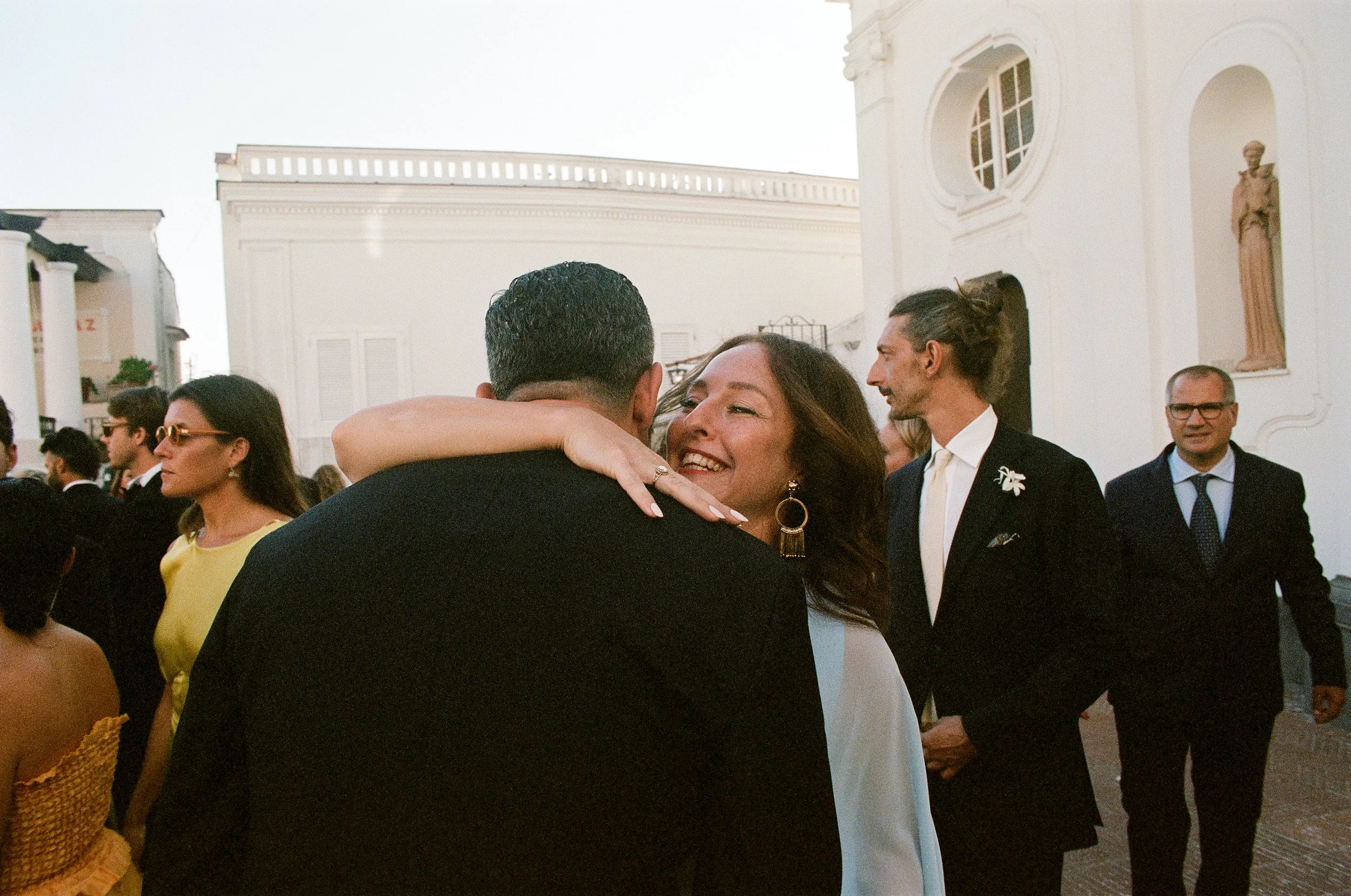 Festa o evento elegante con persone che si abbracciano e sorridono, davanti a un edificio storico o chiesa.