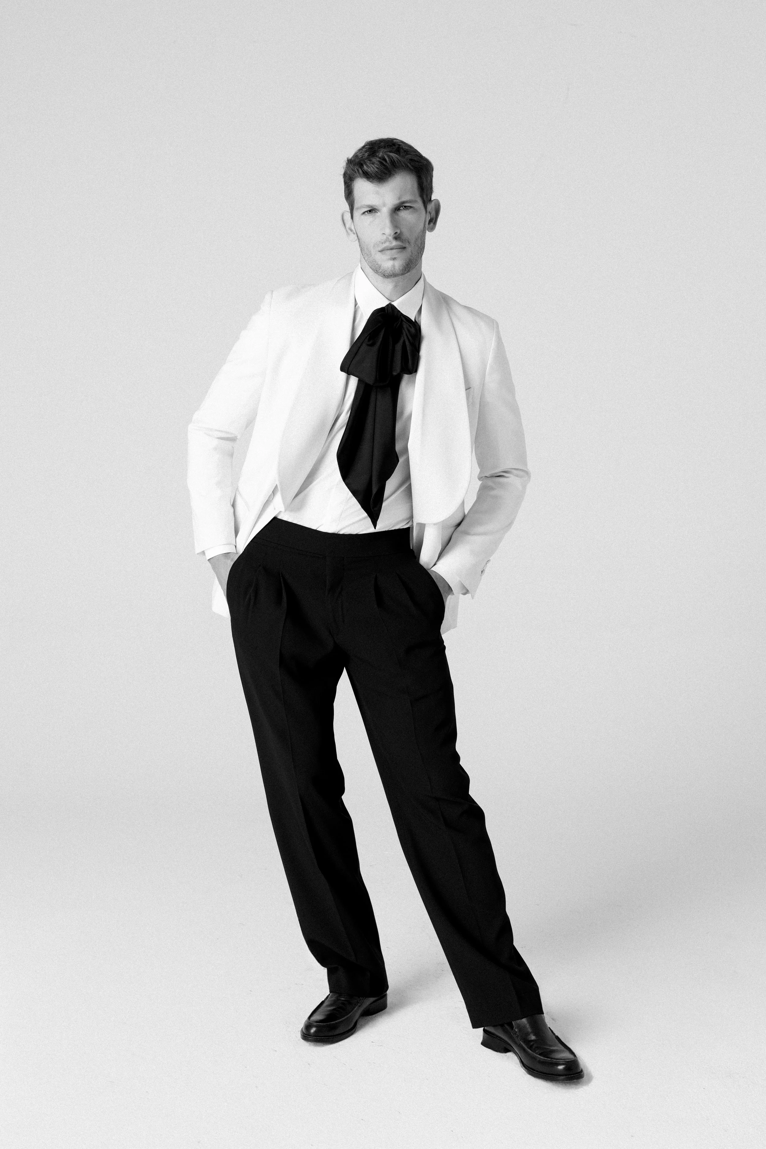 Uomo vestito con cappotto bianco, camicia bianca e cravatta nera, con pantaloni neri e scarpe nere, posando con le mani in tasca.