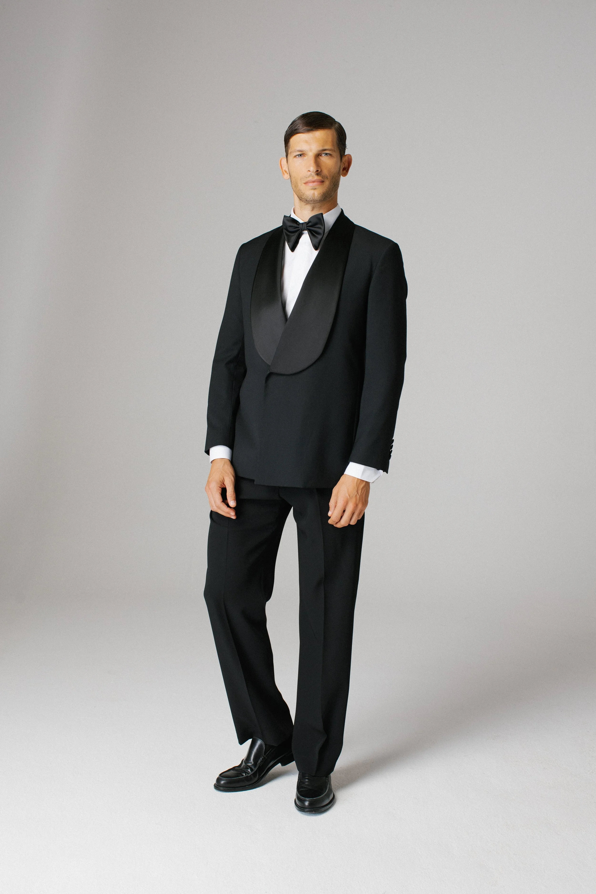 Uomo in Smoking nero con papillon nero, pantaloni neri lucidi e scarpe nere, posa in piedi su uno sfondo neutro.
