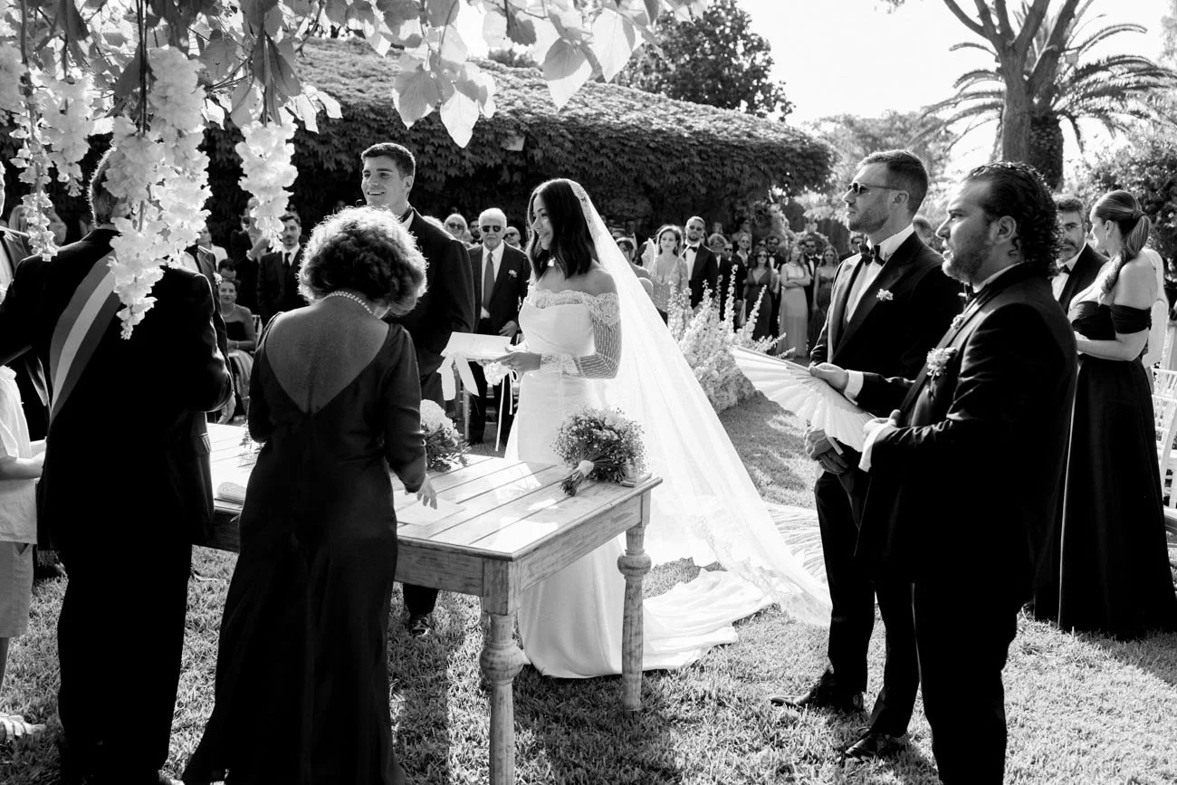 Cerimonia di matrimonio all'aperto con sposi, ufficiali, testimoni e ospiti, ambiente naturale con piante e alberi, foto in bianco e nero.