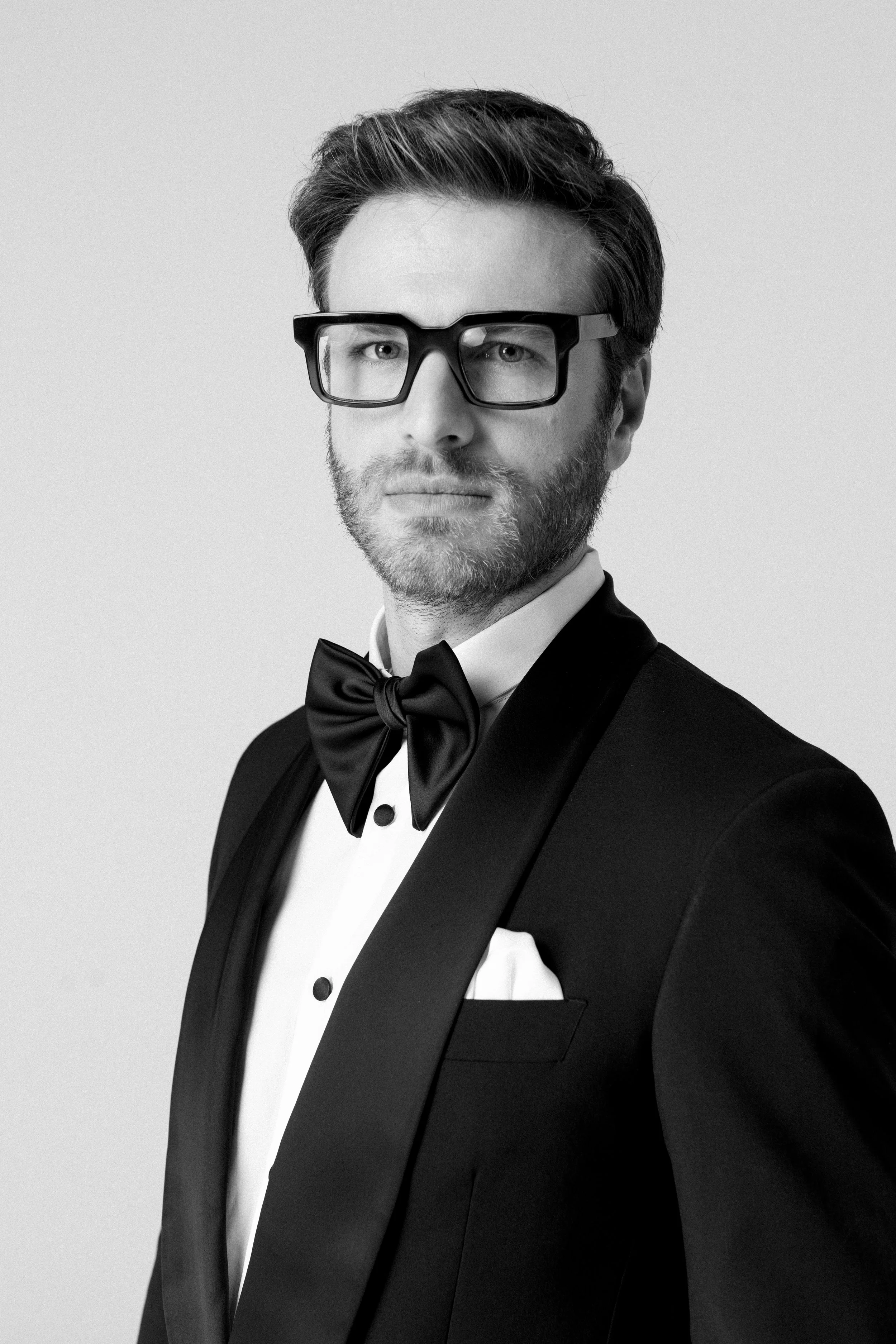 Uomo in abito da sera con papillon nero, occhiali spessi e barba, in bianco e nero, che guarda verso la fotocamera.