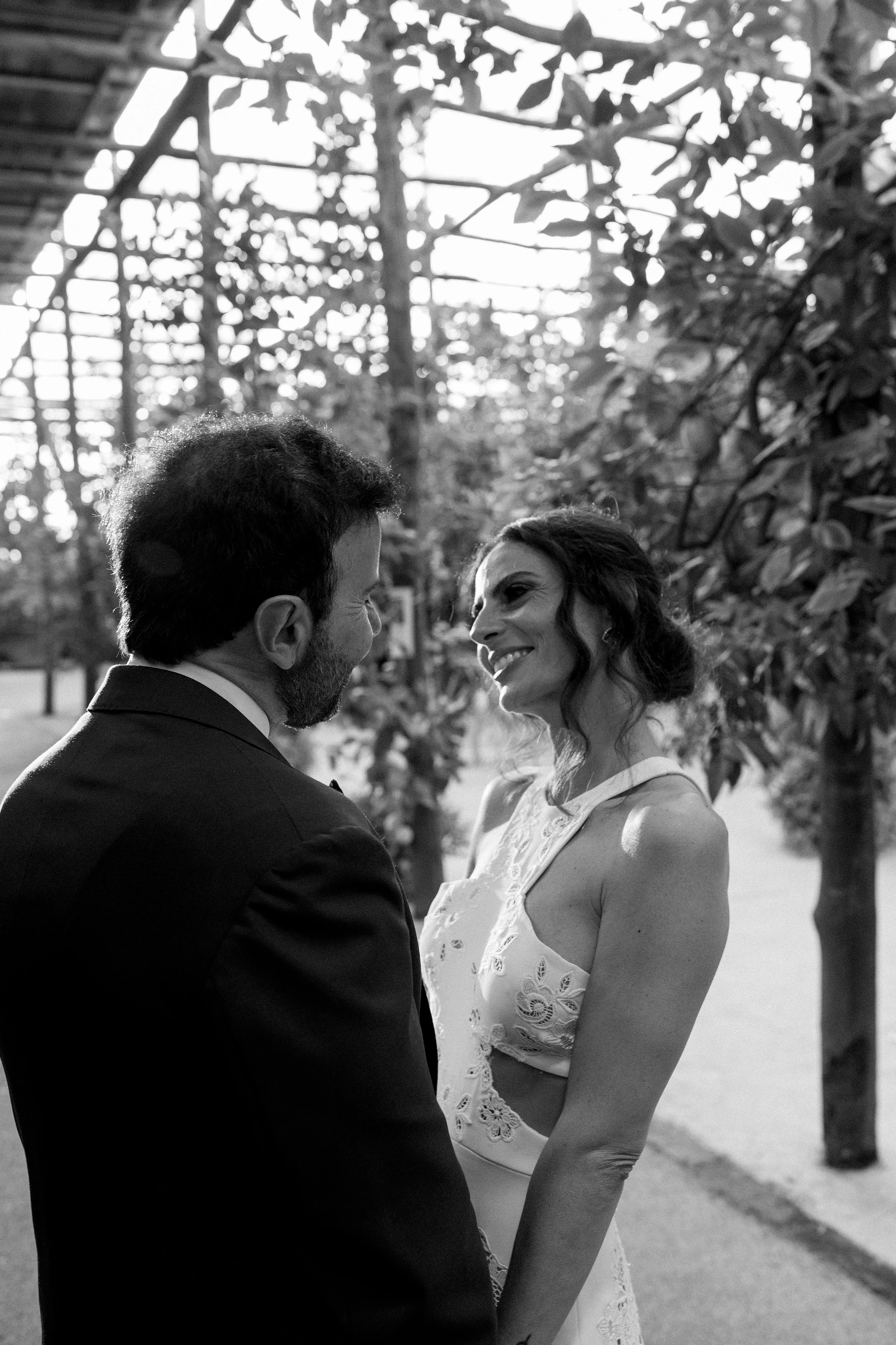 Una coppia sorridente si tiene per mano all'aperto, in un giardino, in un momento intimo e felice, in bianco e nero.