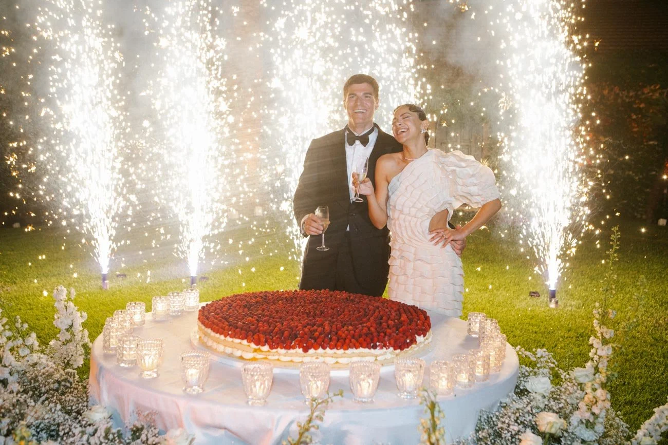 A man and a woman in abito elegante festeggiano un compleanno con una torta di frutti rossi e fuochi d'artificio