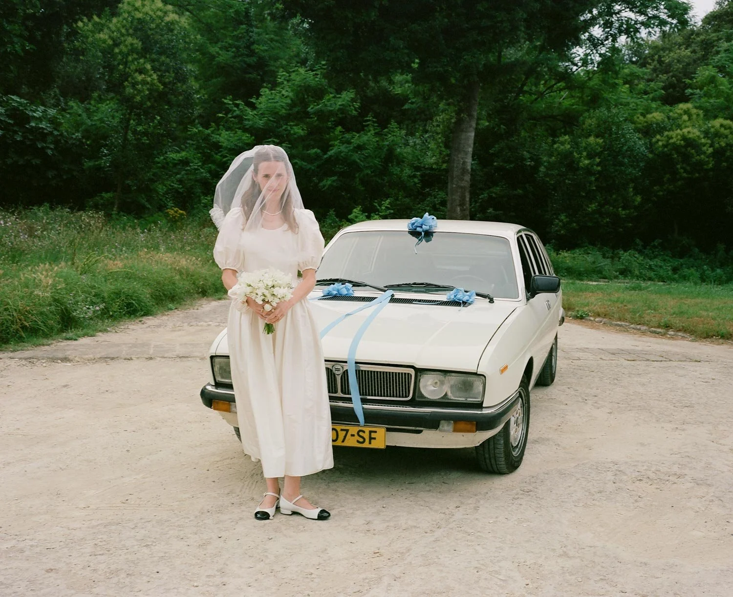 Una donna con un vestito bianco e un velo da sposa tiene un bouquet di fiori davanti a un'auto bianca decorata con fiocchi blu, in un'area aperta con alberi sullo sfondo.