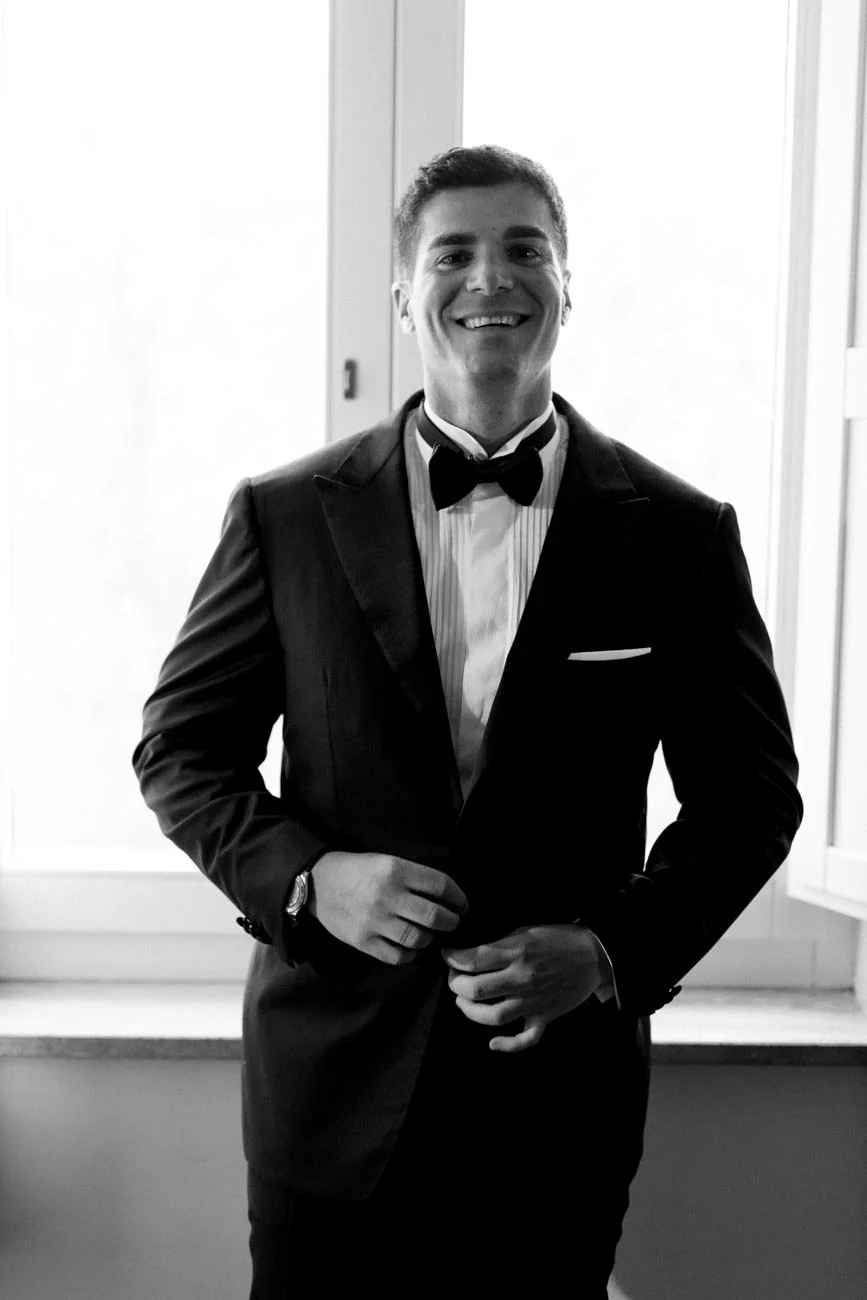 Un uomo sorridente indossa un abito da sera con papillon, in piedi davanti a una finestra, in un ambiente interno.