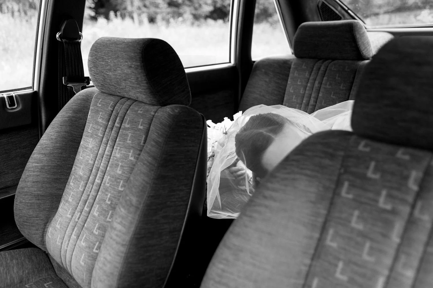 Interno di un'auto con sedili in tessuto e una persona sdraiata con un abito bianco, vista dal lato con finestra che mostra un paesaggio naturale.