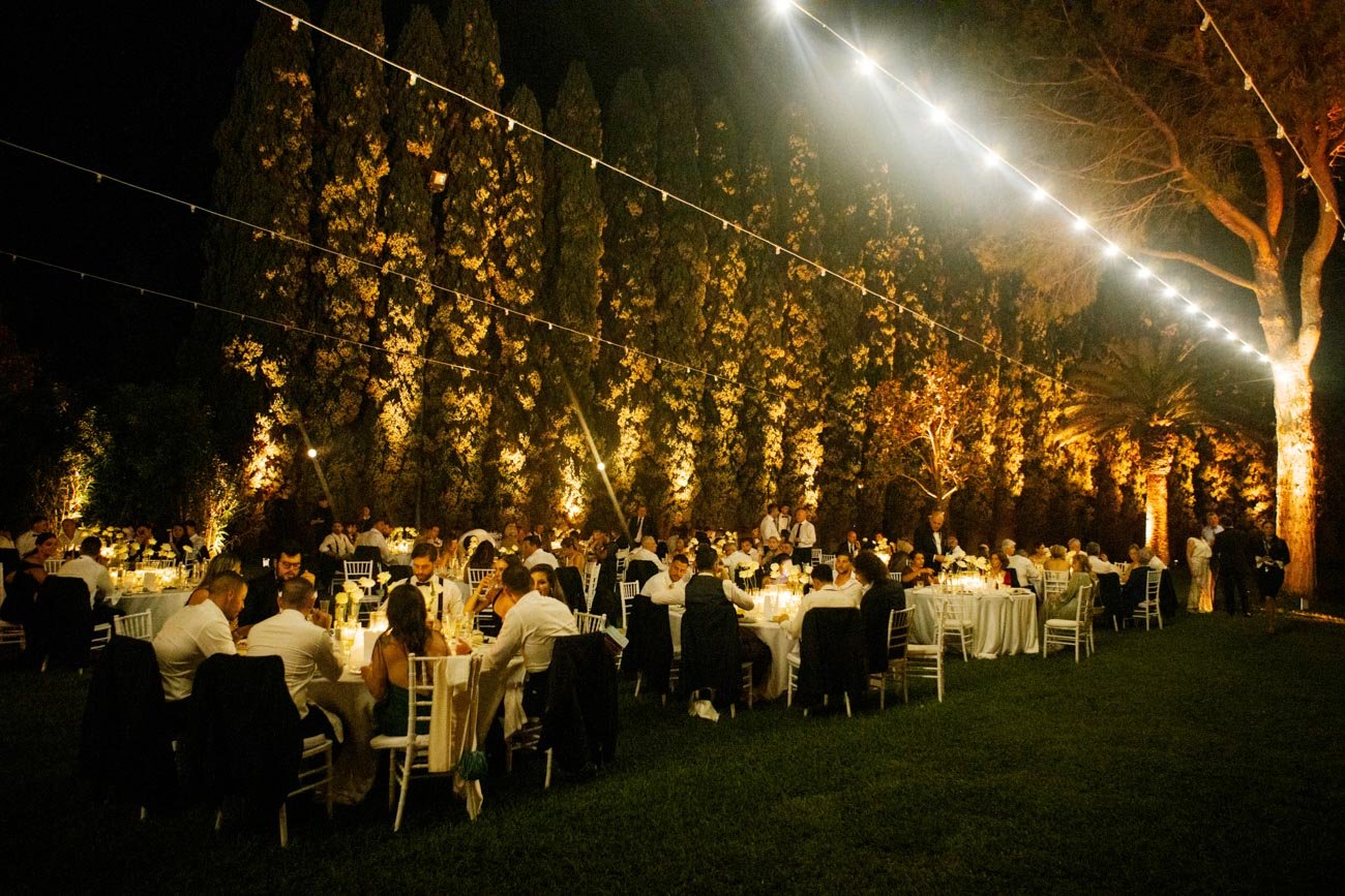Cena all'aperto durante la sera con tavoli e sedie bianche, illuminata da luci sospese, in un giardino con alberi alti e fiori gialli sullo sfondo, con molte persone sedute e che si godono l'evento.