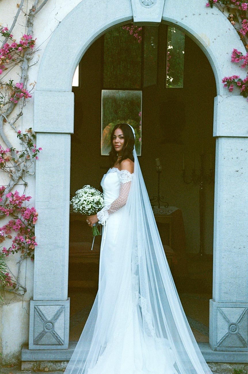 Sposa in abito bianco con bouquet di fiori bianchi, posa in un arco decorato con fiori rosa.
