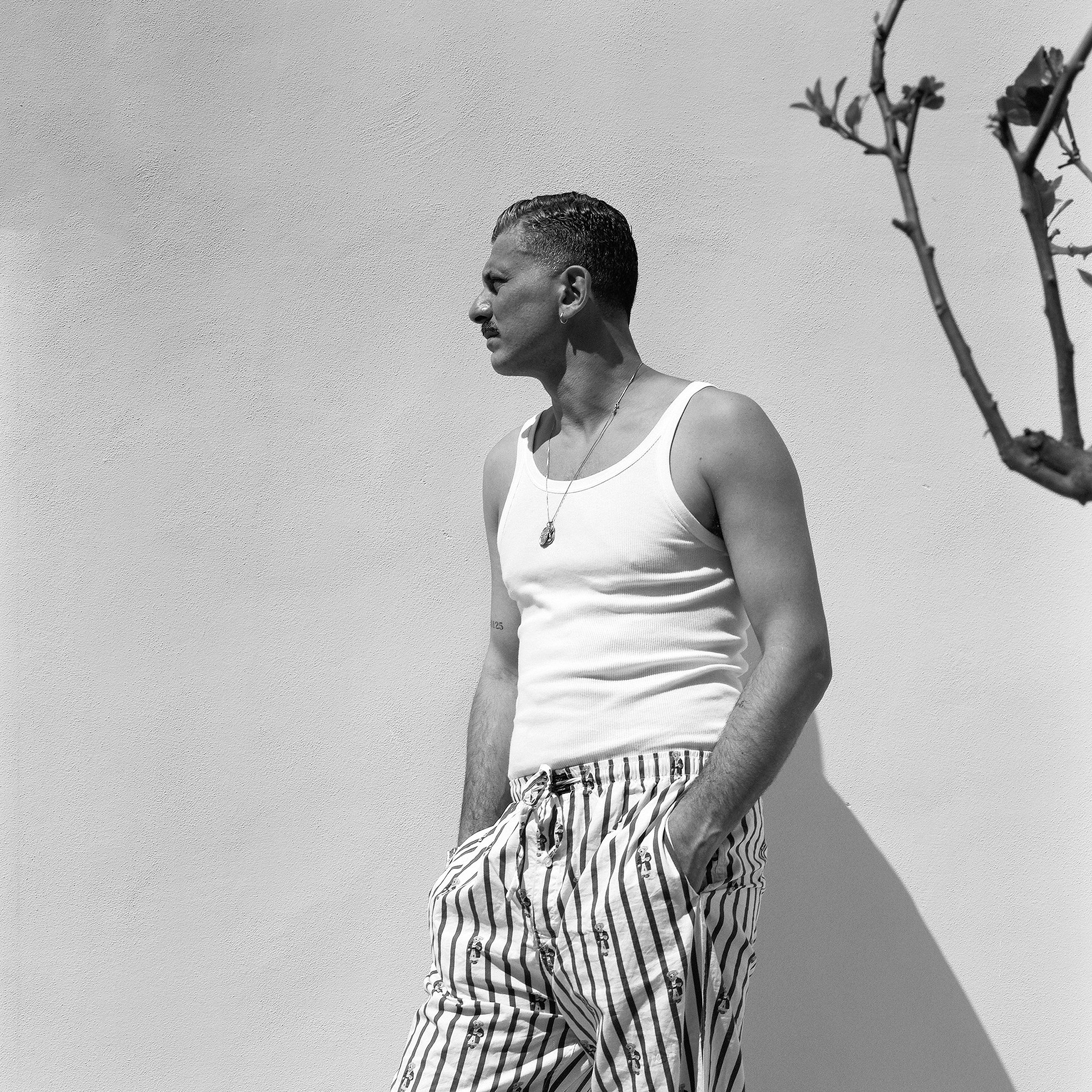 Uomo con camicia a righe e pantaloni a righe, in piedi contro una parete bianca, in posizione di profilo, con mani nelle tasche, in una scena in bianco e nero.