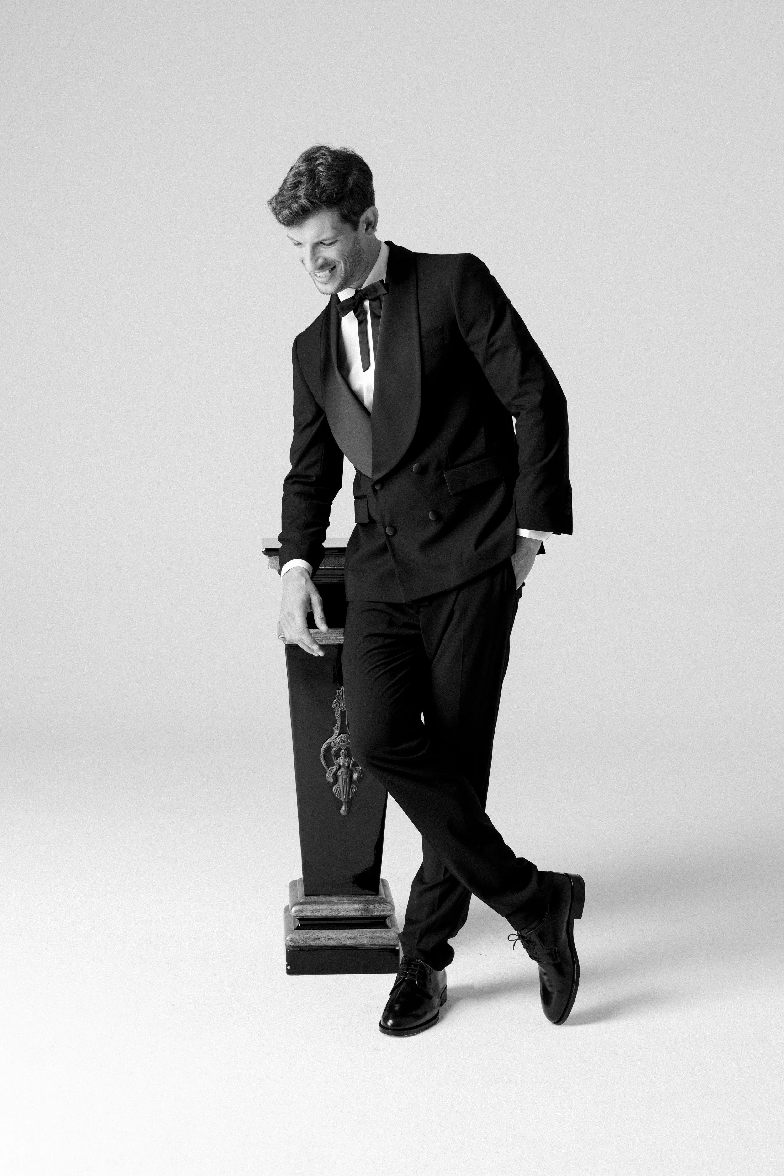 Uomo in abito elegante nero, in posa con sorriso, accanto a un complemento decorativo.