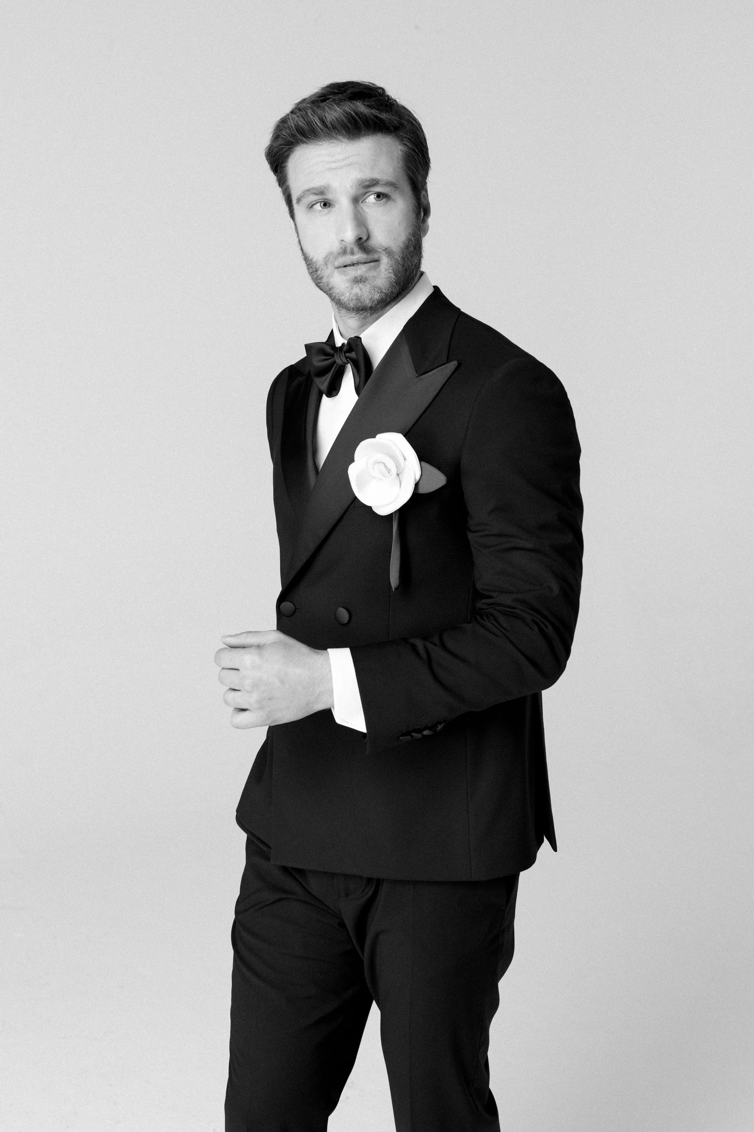 Un uomo in abito da sera nero con papillon e una rosa bianca sulla giacca, posa con un’aria seria in uno studio fotografico.