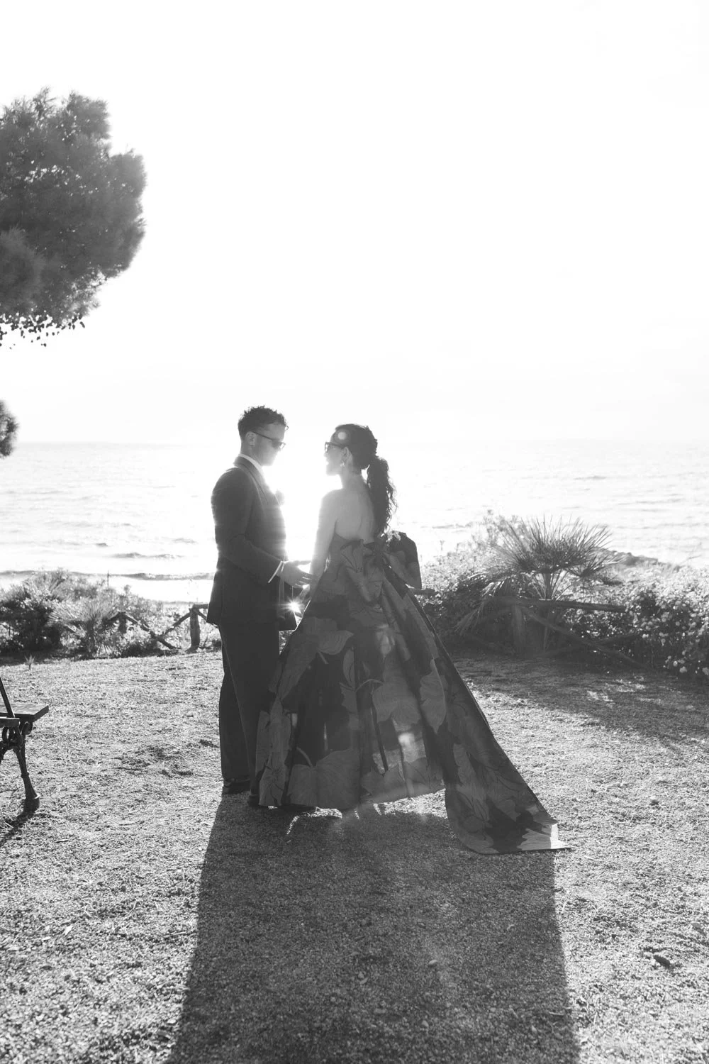 Una coppia di persone vestite elegantemente, presumibilmente sposi, si tiene per mano sulla spiaggia al tramonto, con l'oceano sullo sfondo.