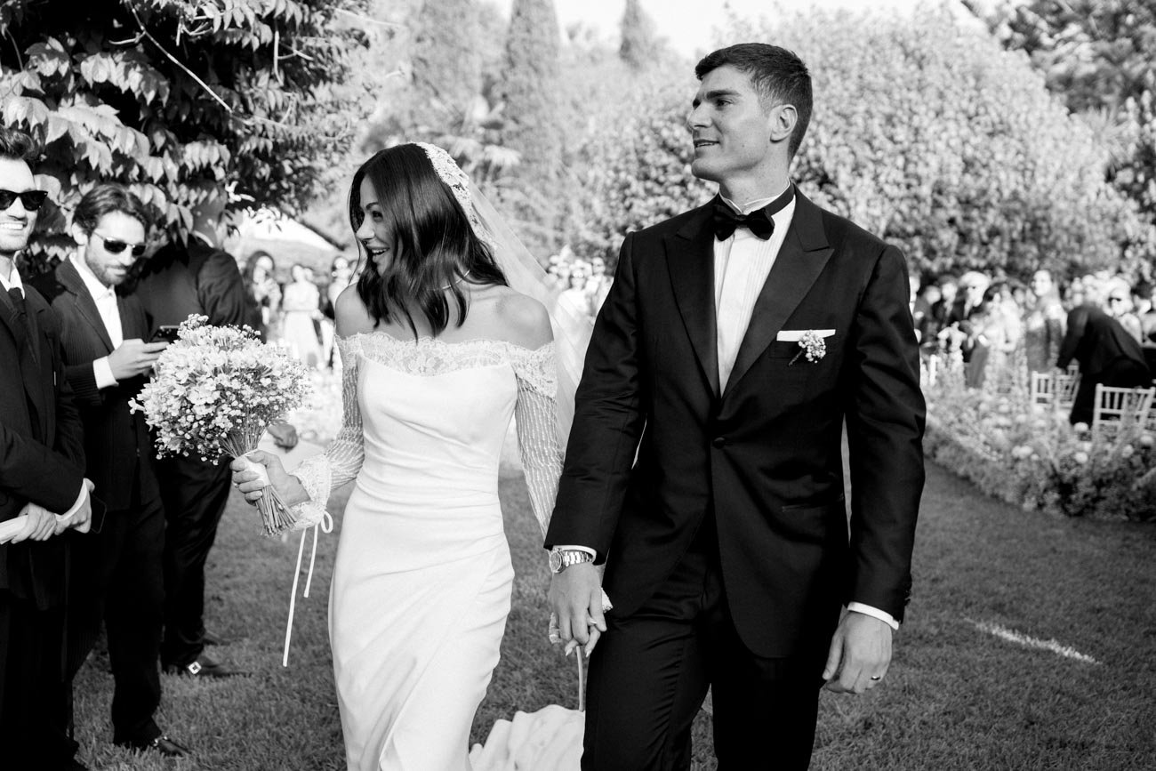 Una coppia sposa e sposo camminano insieme durante un matrimonio all'aperto, circondati da invitati, alberi e decorazioni floreali.