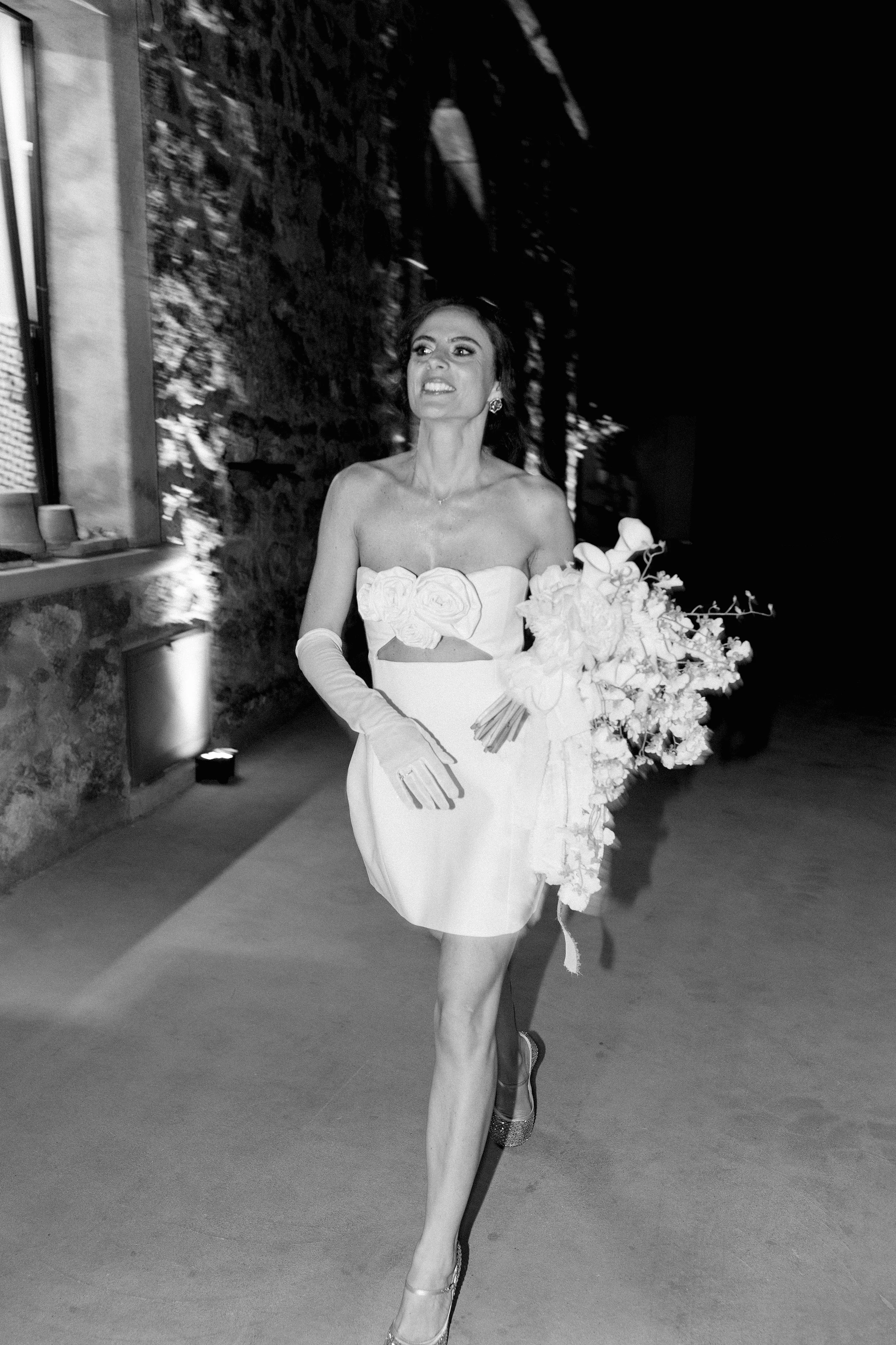Una donna in abito da sposa con un grande bouquet di fiori, cammina sorridente in un ambiente con pareti di pietra e illuminazione soffusa.