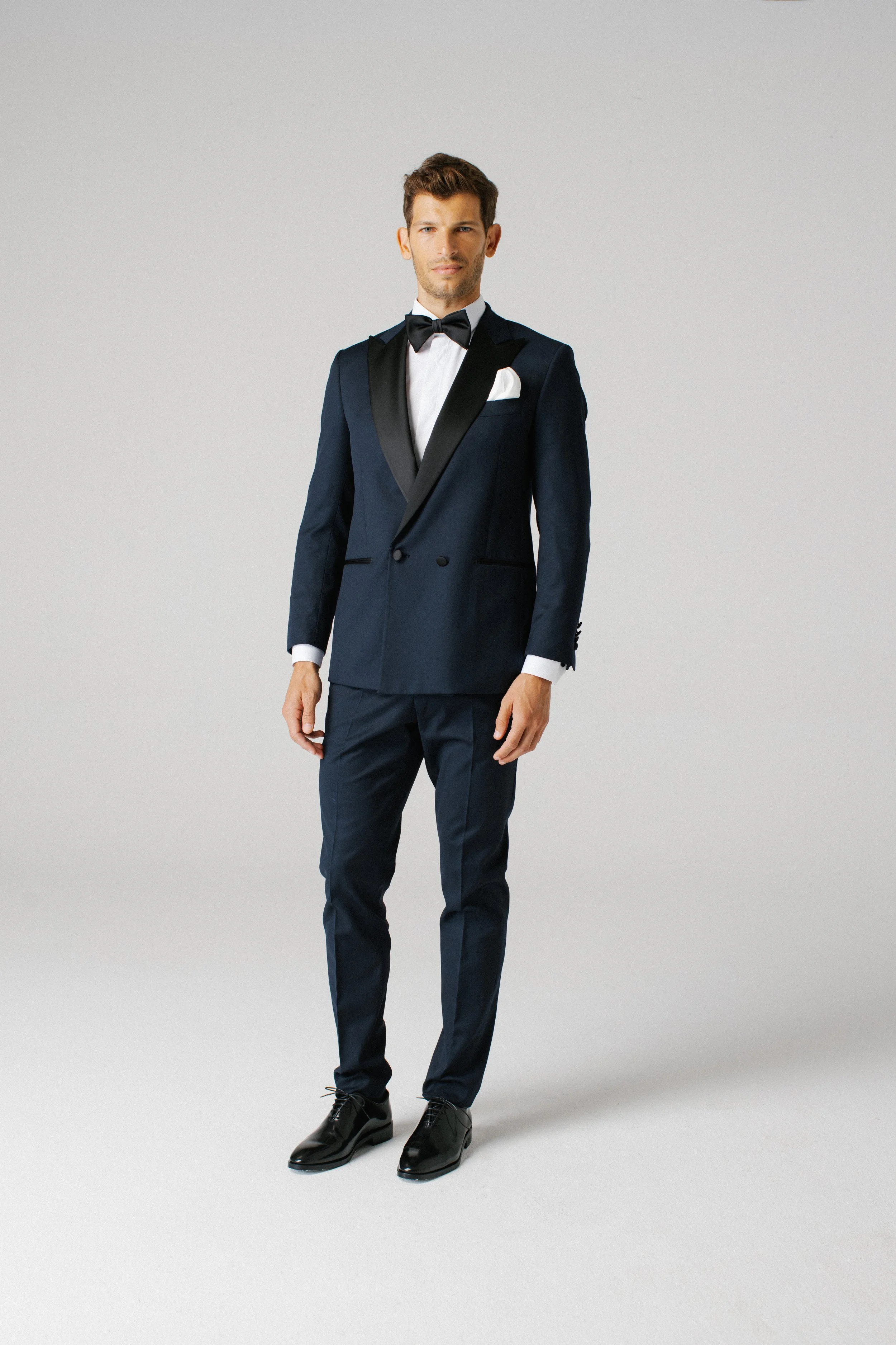 Uomo vestito con abito blu scuro con revers in seta nera, camicia bianca e cravatta a farfalla nera, in piedi davanti a uno sfondo grigio chiaro.