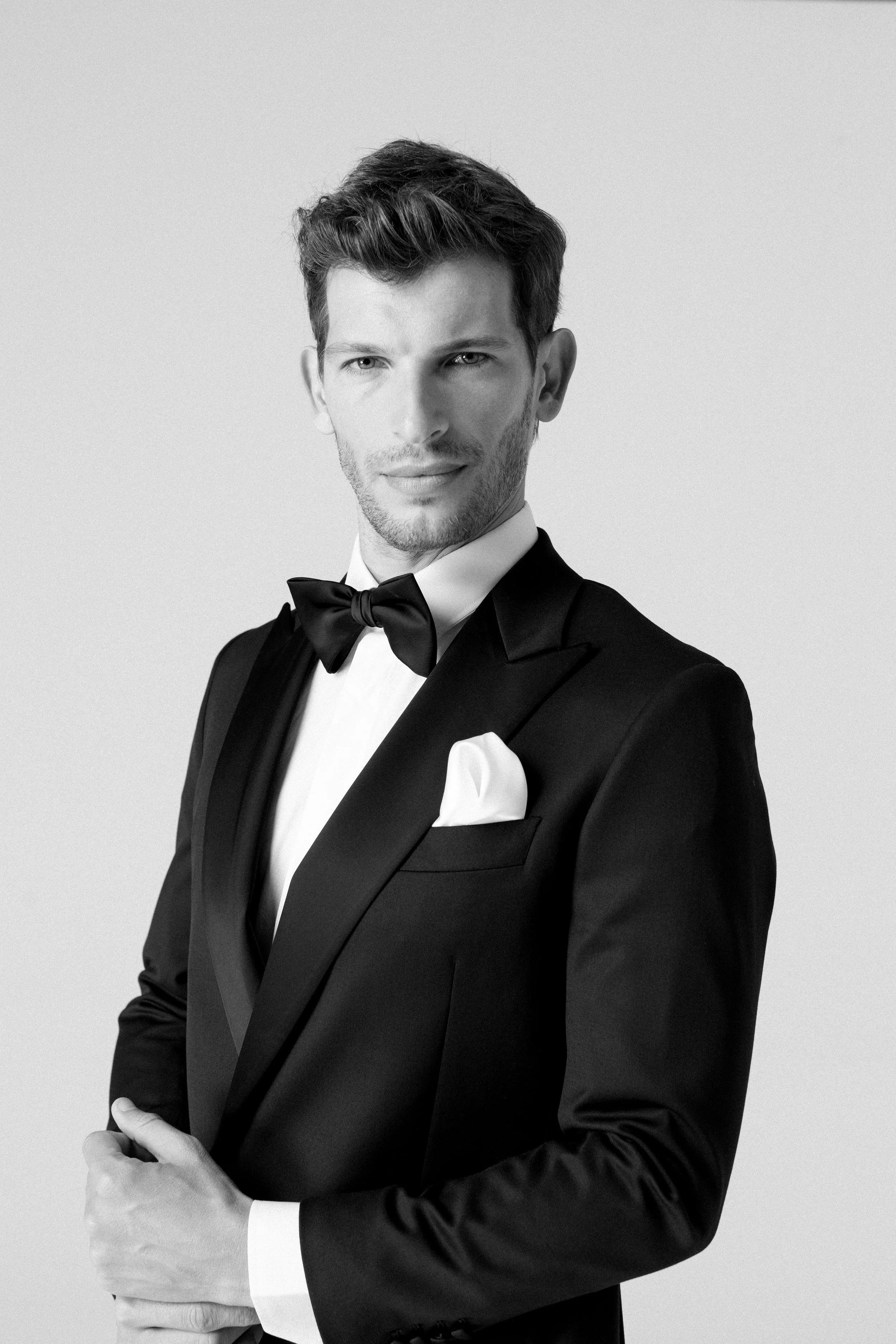 Uomo in abito da sera nero con papillon e pochette bianca, sfondo neutro.