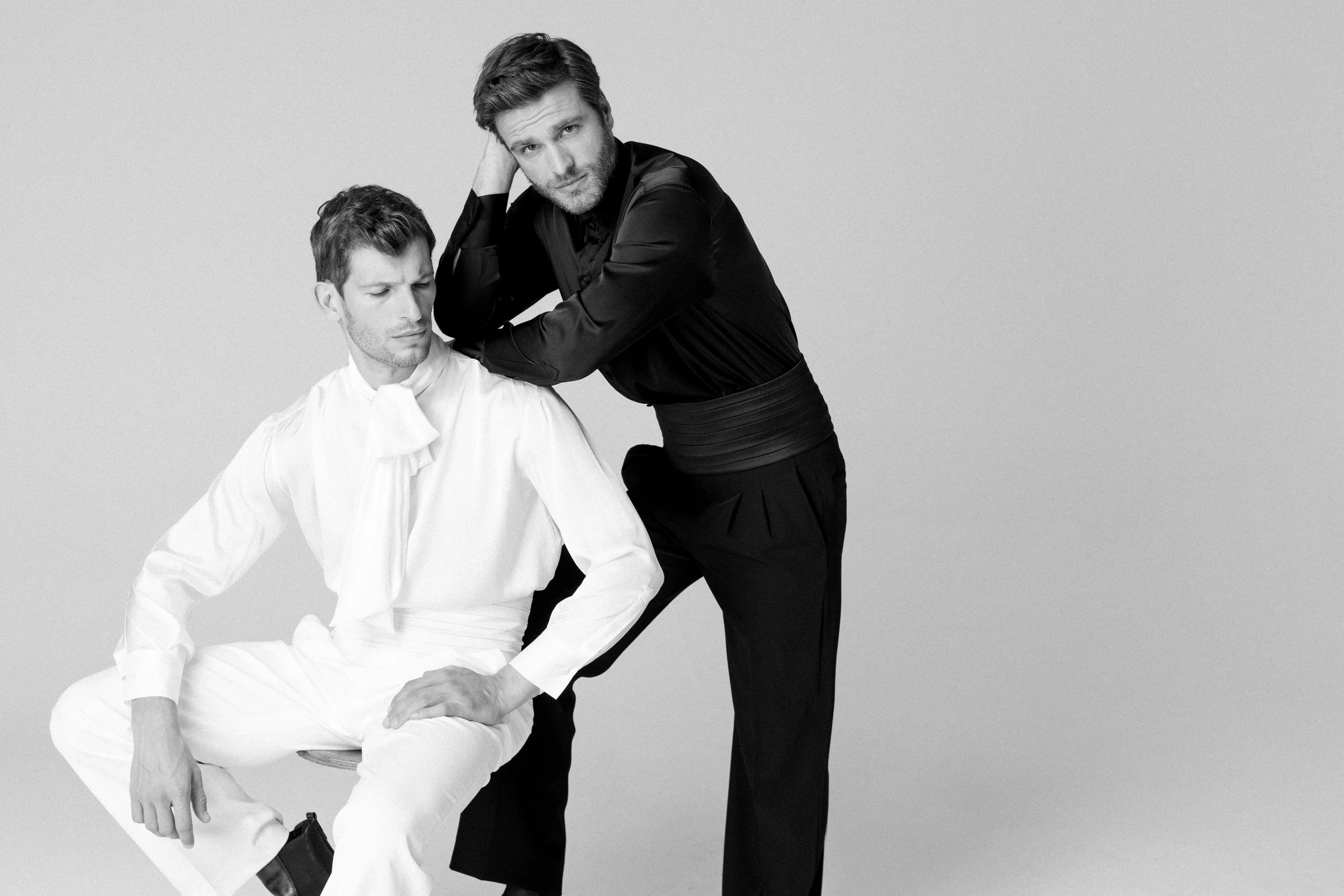 Due uomini in abiti eleganti, uno seduto con abito bianco e uno in piedi con abito nero, posano per una foto in bianco e nero