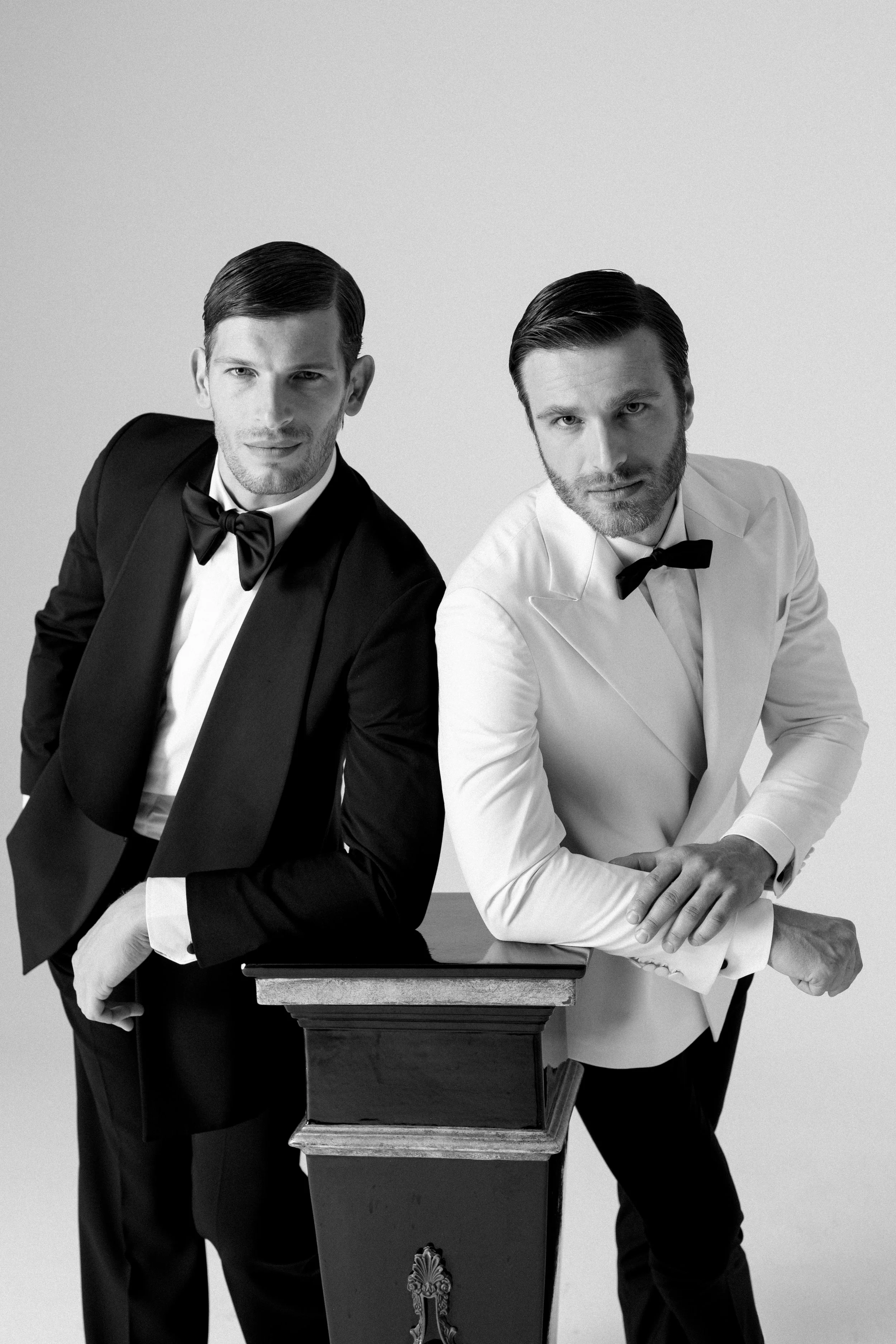 Due uomini in abiti formali, uno in smoking nero e l'altro in giacca bianca, con sfondo semplice, in una fotografia black e white.