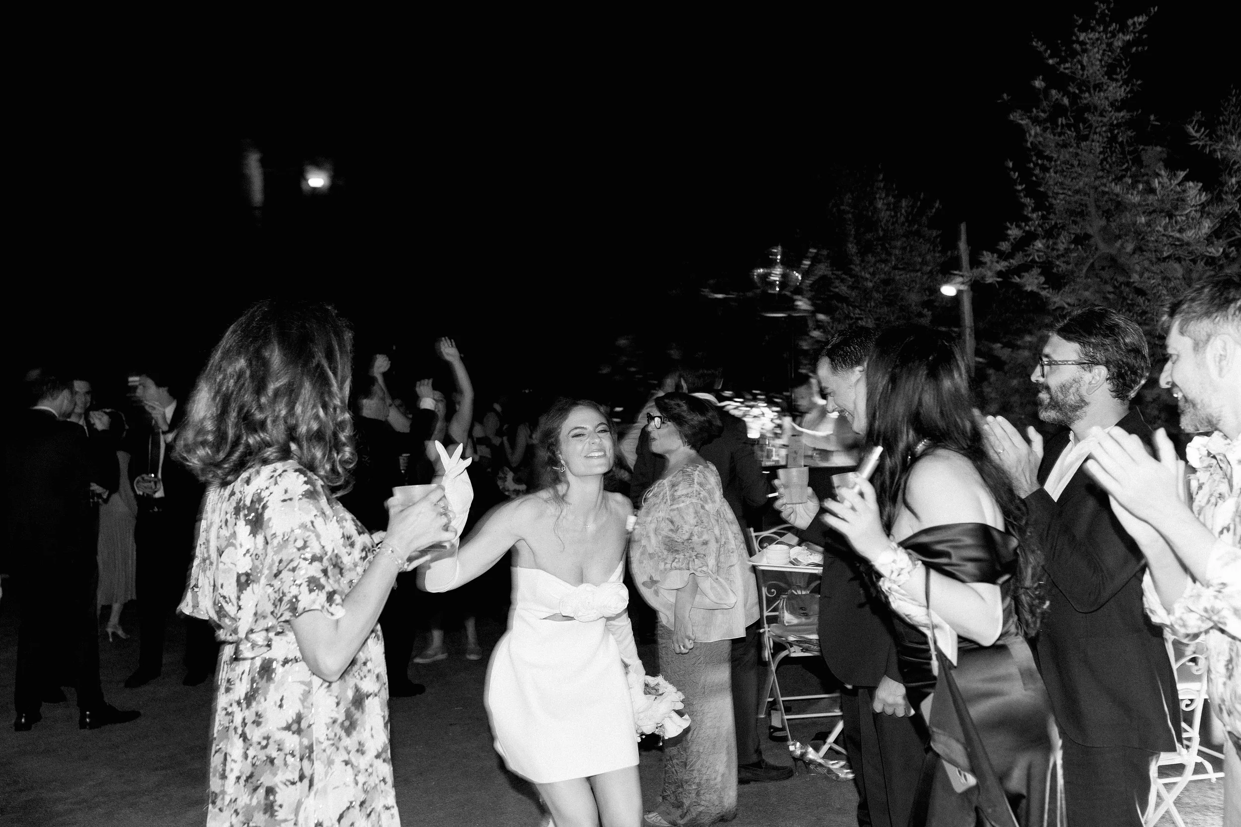 Una scena di festa notturna con persone che ballano, bevono e si divertono all'aperto, con alcune persone che applaudono e altre che si stanno abbracciando o scambiando auguri.