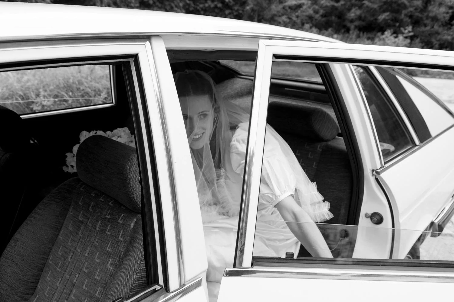 Sposa seduta sull'auto, visibile dal parabrezza, con sorriso, indossa un vestito bianco e un velo, mentre guarda fuori dal finestrino.