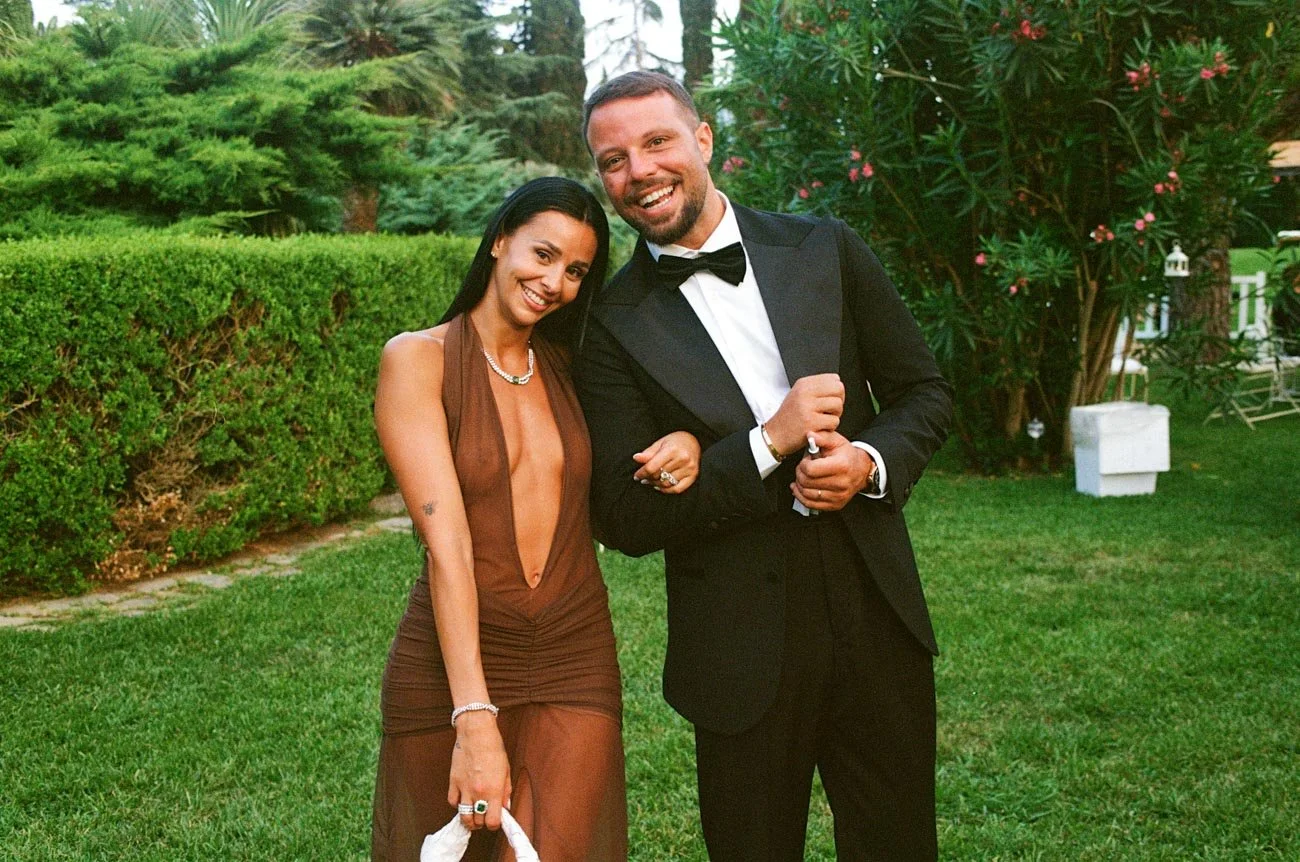 Una coppia elegante sorridente in un giardino verde, con uomini in abito nero con papillon e donna in vestito marrone senza schiena.