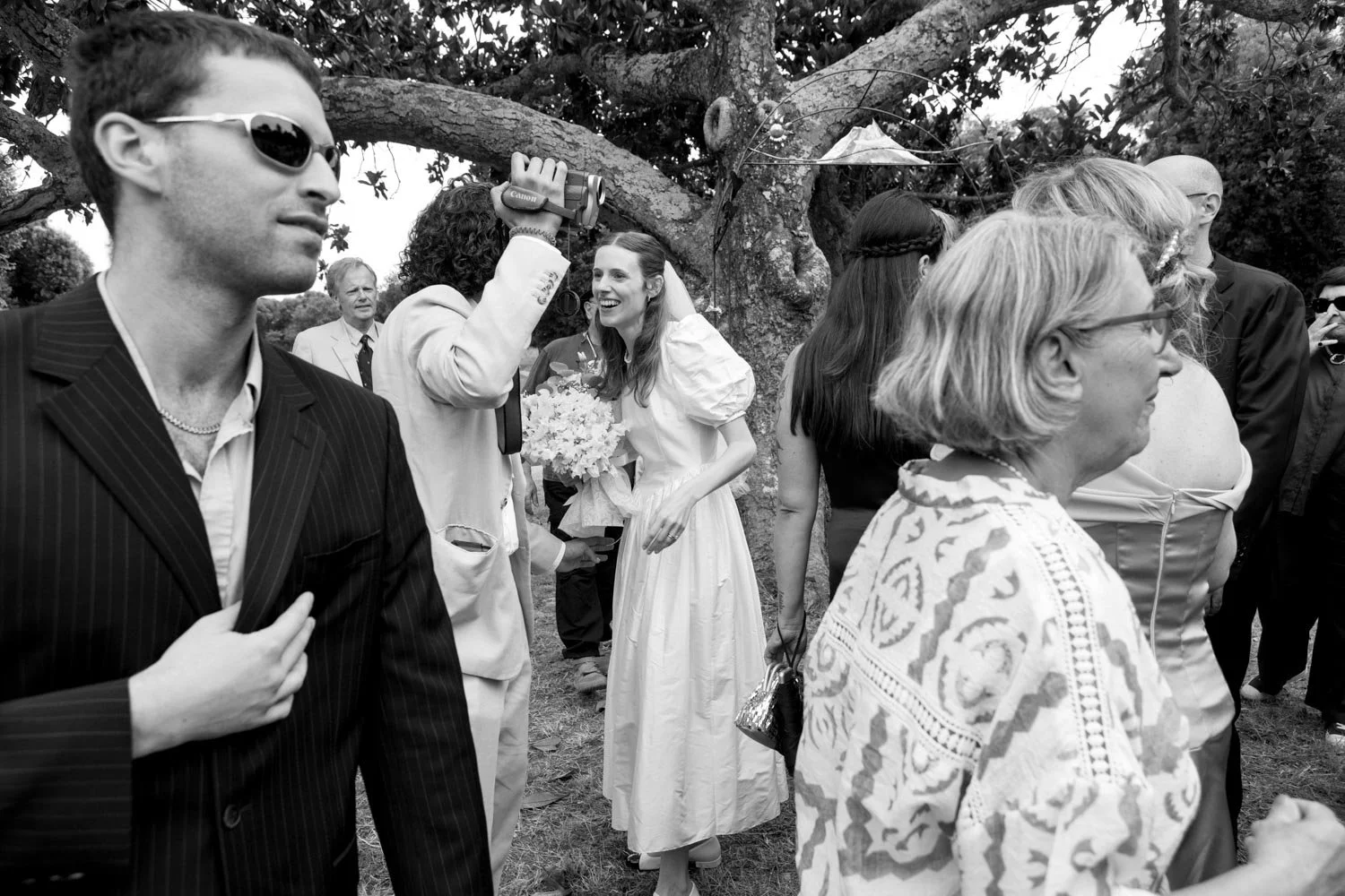 Una scena di un matrimonio con molte persone intorno, una sposa sorridente con un mazzo di fiori in mano, circondata da amici e parenti, sotto un albero.
