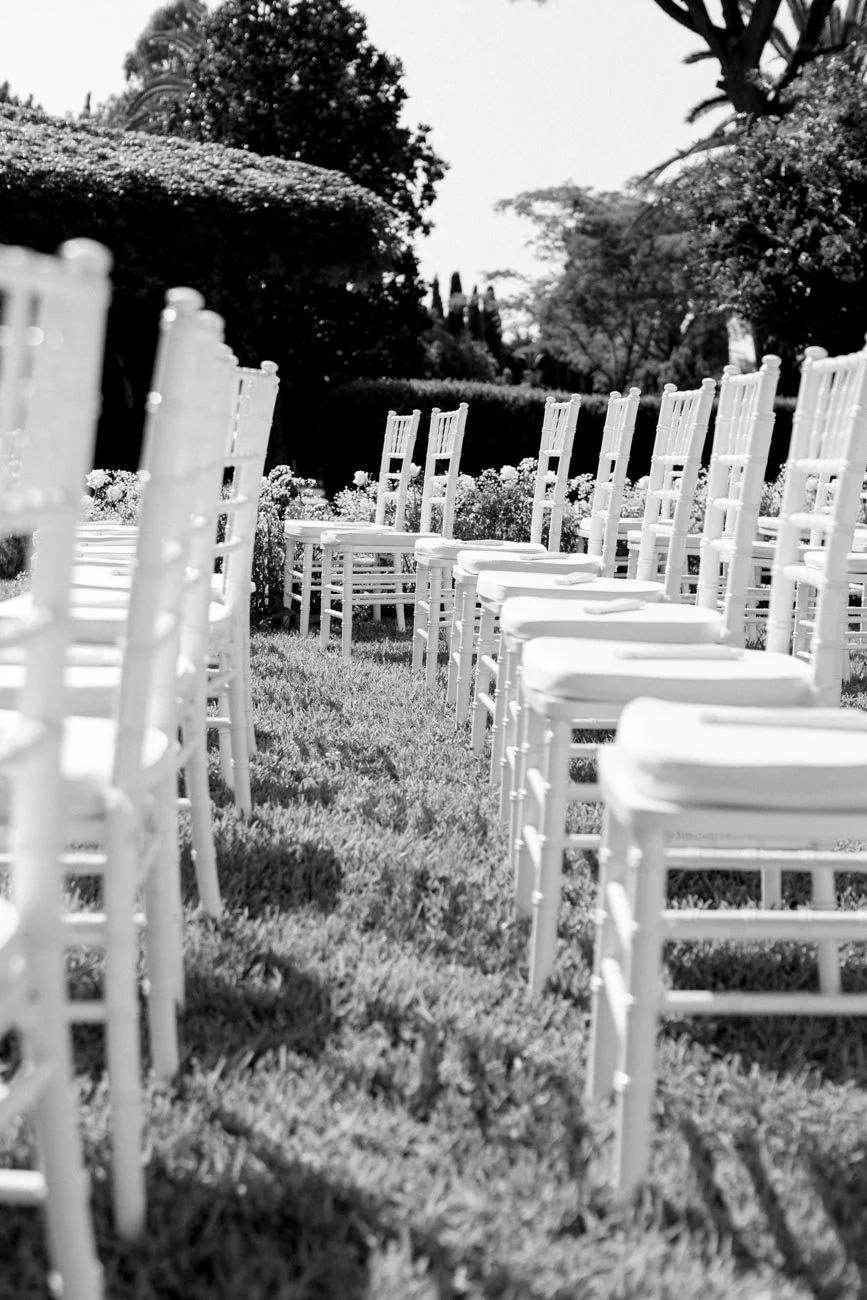 Sedie bianche disposte in fila su un prato in un giardino, con piante e alberi sullo sfondo.