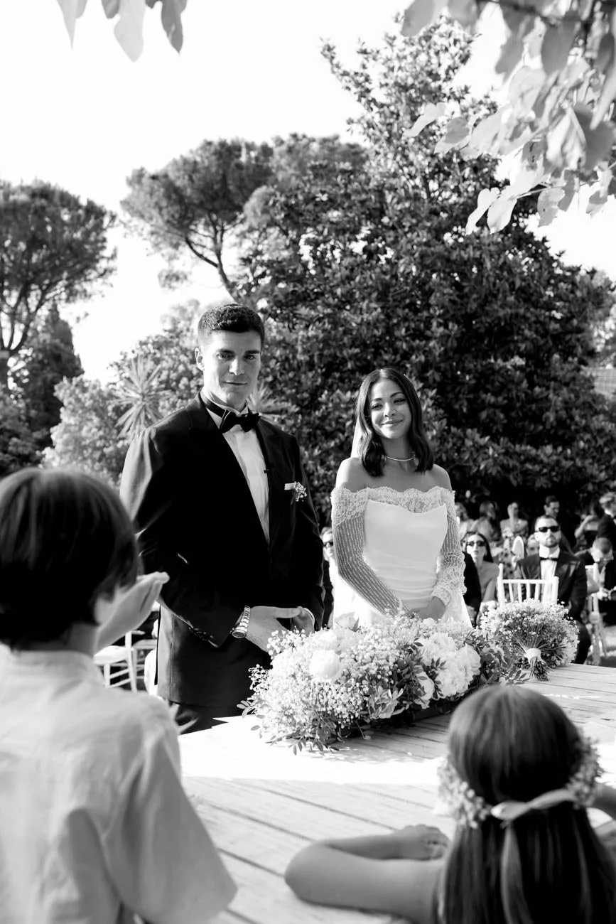 Cerimonia di matrimonio all'aperto con una coppia di sposi, un uomo in smoking e una donna in abito da sposa, circondati da amici e familiari, con alberi sullo sfondo.