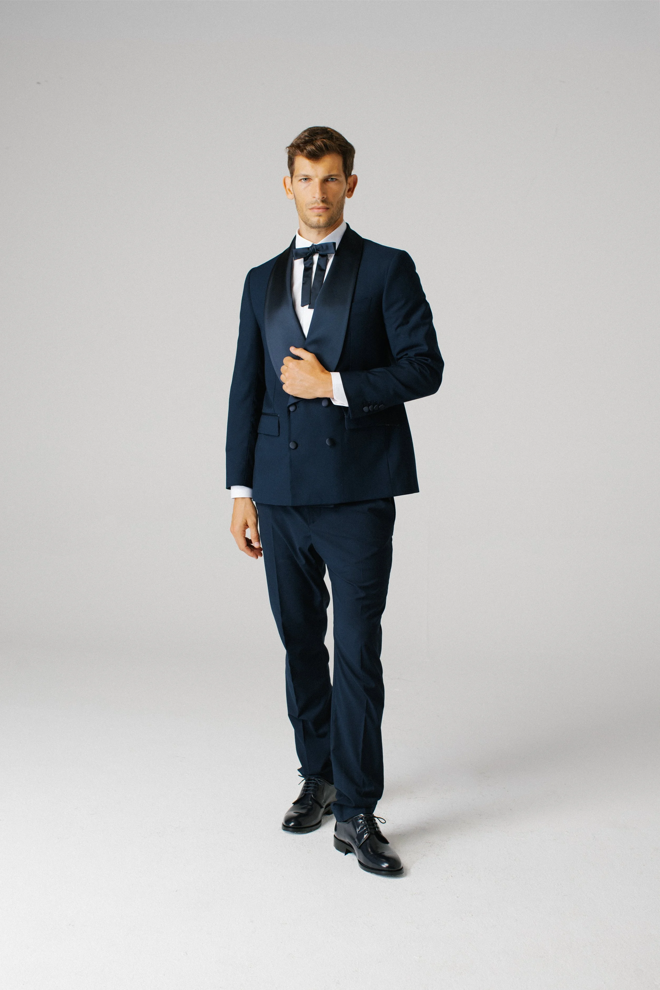 Uomo che indossa un abito da sera blu scuro con papillon, in piedi su uno sfondo neutro.