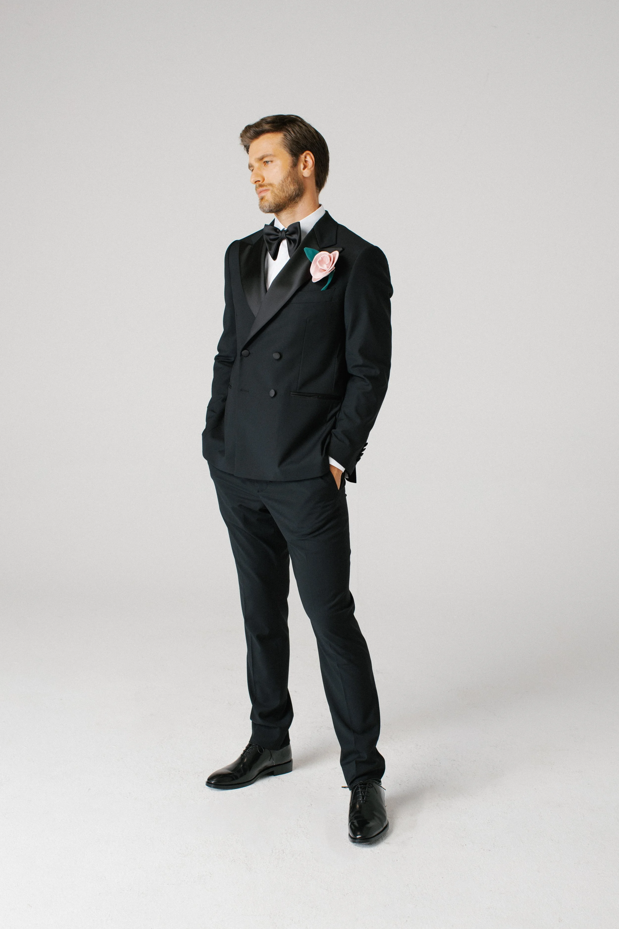 Uomo in abito da sera nero con papillon nero, taschino con rosa e faccia seria che guarda a sinistra