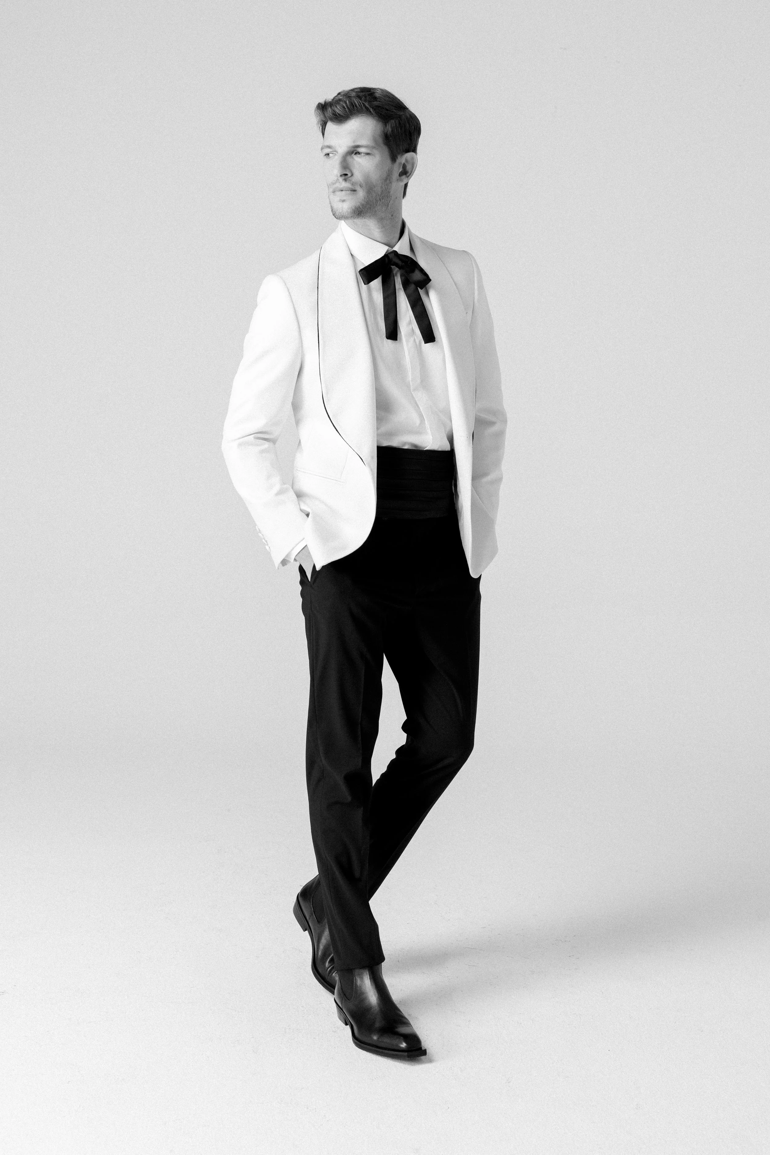 Uomo in abbigliamento elegante, con giacca bianca, pantaloni neri e scarpe nere, posa con le mani nelle tasche, in un setting neutro.