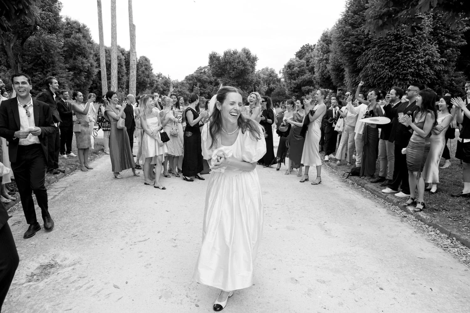 A woman in a white dress walks down a path, smiling, surrounded by a crowd of well-dressed people clapping and taking pictures, outdoors with trees on both sides.