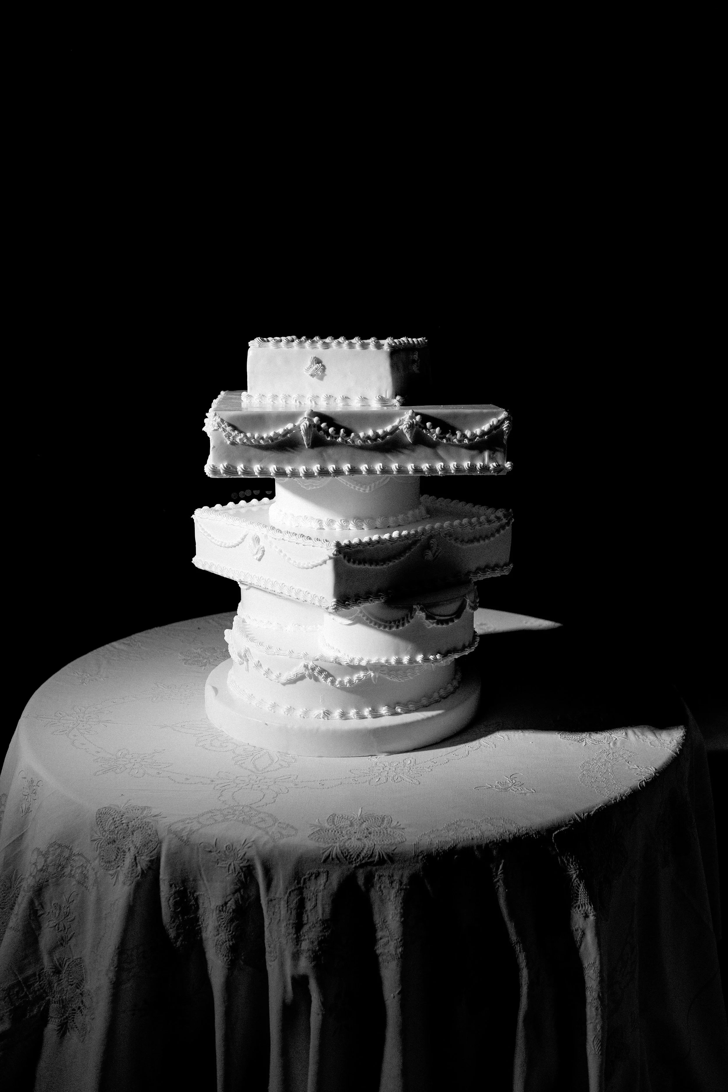 Torta nuziale a più livelli decorata con ricami e nappe, posizionata su un tavolo con tovaglia semplice, ambientata in un ambiente con luce mirata e sfondo nero.