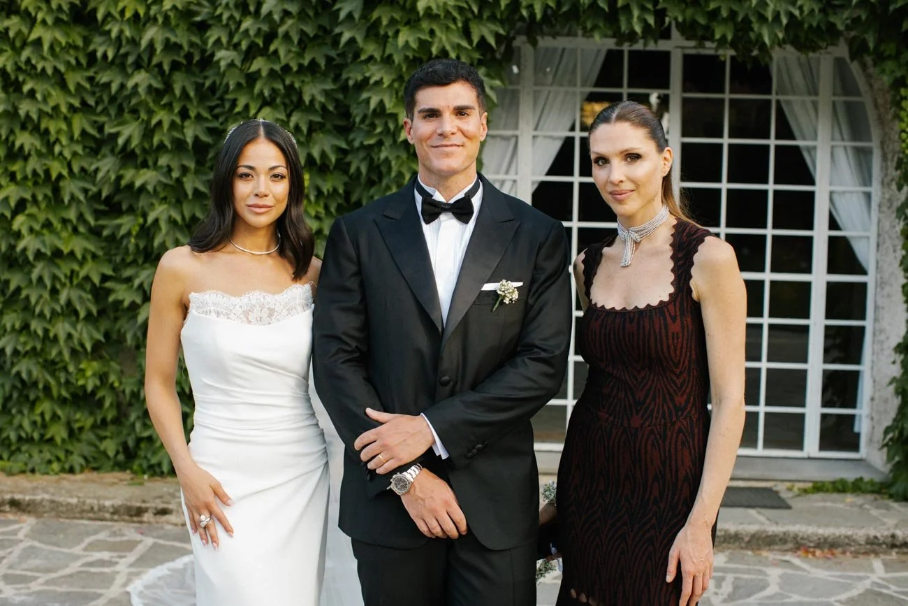 Tre persone in abiti eleganti, un uomo in completo scuro con papillon, una donna in abito bianco da sposa e un'altra donna in vestito nero con stampa rossa, si trovano davanti a una casa con pareti di edera verde. È un matrimonio.